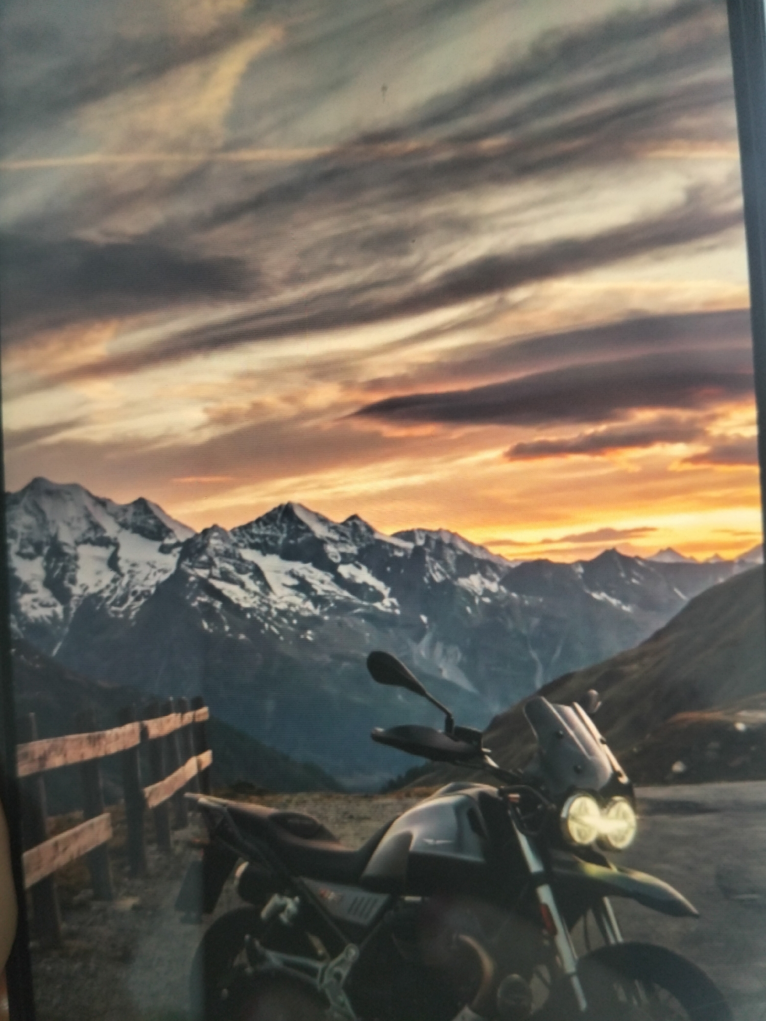 A dramatic landscape photograph depicts a dirt track in a mountainous region, likely at sunset. The sky is filled with streaks of orange, yellow, and gray clouds, suggesting the sun is low on the horizon. In the mid-ground, snow-capped mountains rise majestically against the vibrant sky. In the foreground, a dual-sport motorcycle is parked on the dirt track, facing towards the viewer. The motorcycle has a prominent windshield, twin headlights, and appears to be dark in color. To the left of the motorcycle, a wooden fence runs along the side of the track, dividing it from the rugged terrain. The scene is devoid of any people or animals. The overall impression is one of rugged beauty and solitude. There is no indication of the city of Payagaboh, Indonesia in this image.