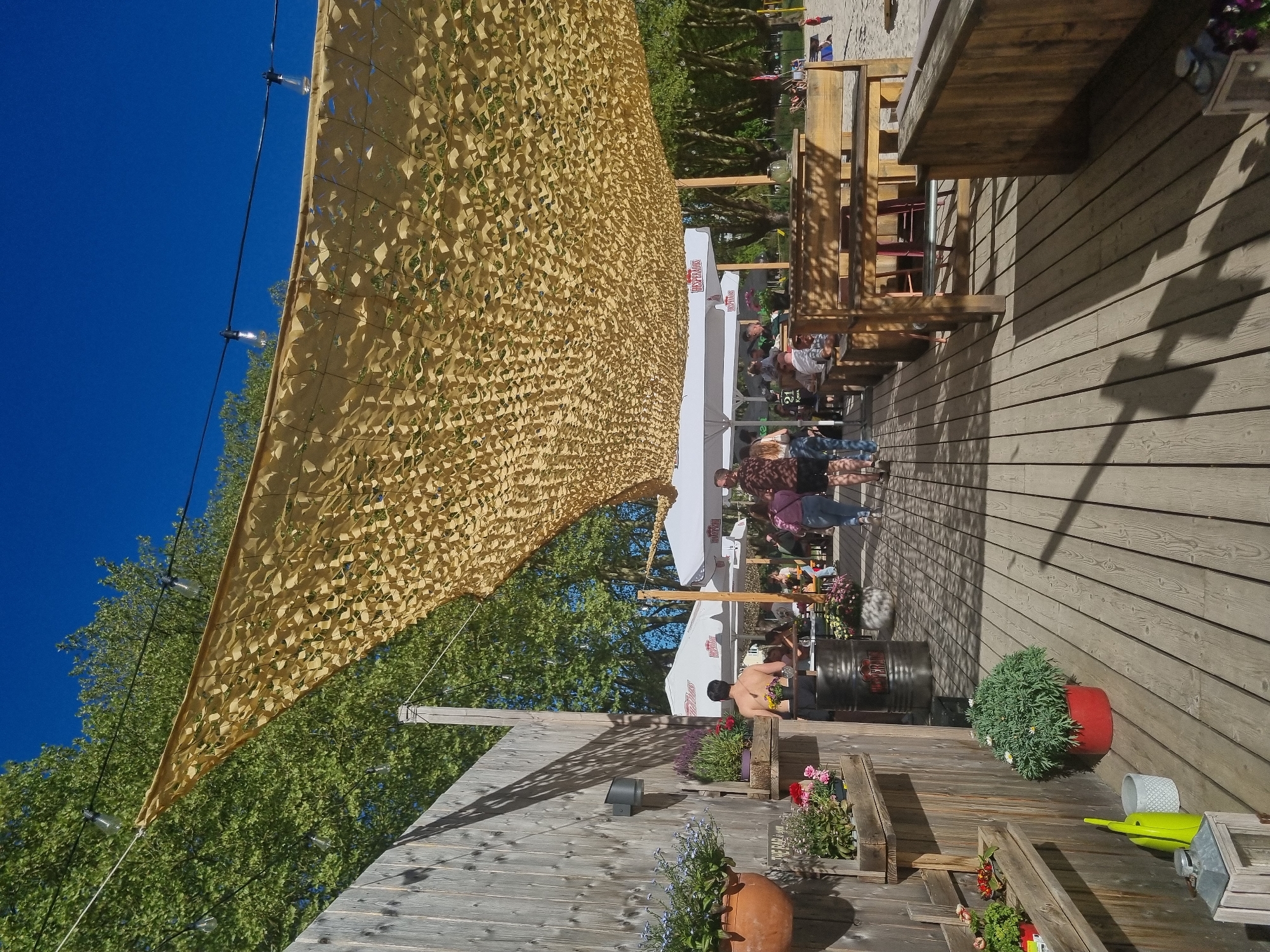 The image depicts an outdoor seating area, potentially a restaurant or bar, located in Saarbrücken, Germany. The scene is illuminated by sunlight filtering through a camouflage net suspended overhead. A wooden deck serves as the floor. People are seen seated at tables under white umbrellas. Planters with flowers are positioned along the wooden wall of the deck. A black barrel-shaped object is also present. The sky is clear and blue.