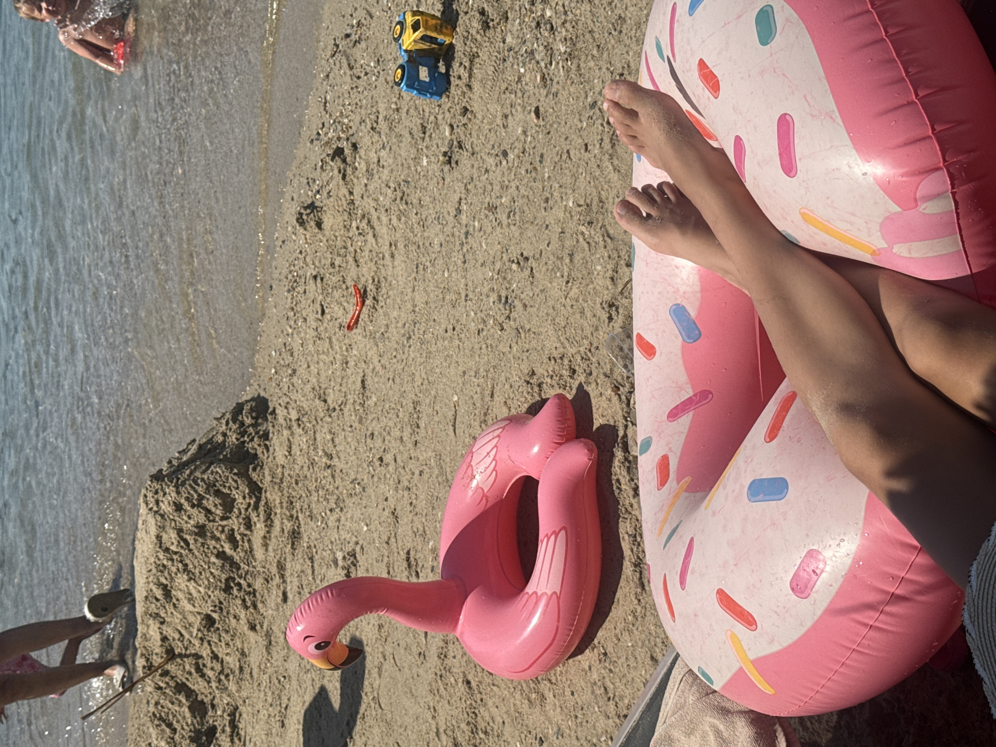 An overhead shot shows a sunny beach in Bodrum, Turkey. In the foreground, a person's bare legs and feet are propped up on a pink inflatable donut pool float. The float is decorated with colorful sprinkles. Behind the float, a pink inflatable flamingo sits on the sand. Further back, a sandcastle stands near the water's edge. In the shallow water, a child is splashing and playing. The scene captures a relaxed summer day at the beach.