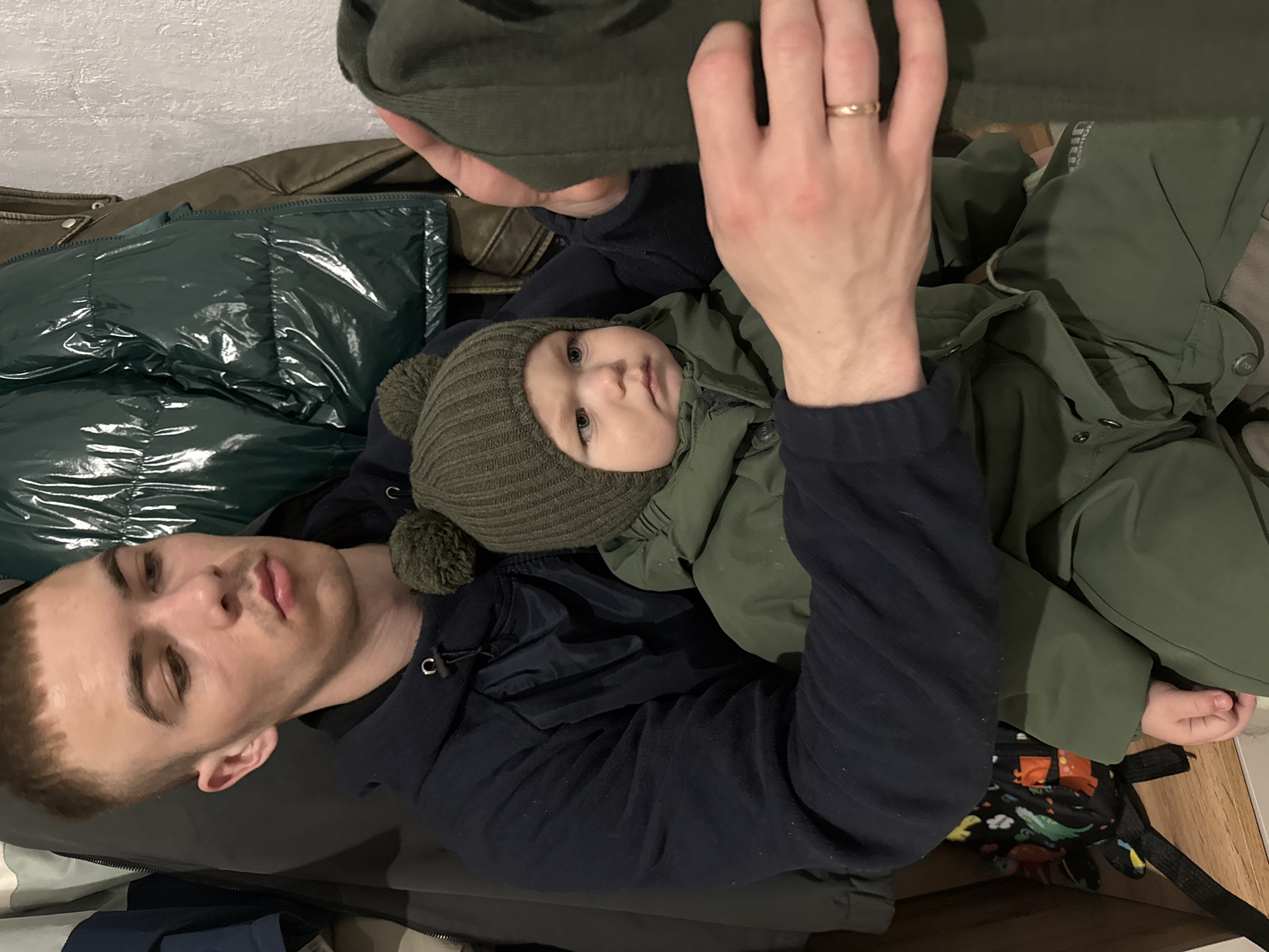 The image shows a man and a baby in an indoor, domestic setting, likely in Chaikovsky, Russia. The man, with light skin, short light-brown hair, and light stubble, lies on his back or side, looking calmly towards the camera. He wears a dark blue long-sleeved hooded top, and a gold wedding ring is visible on the ring finger of his left hand, which is raised.
The baby is nestled on the man's chest or lap, also looking towards the camera with a neutral to slightly curious expression. The baby has light skin, blue eyes, and is dressed warmly in a dark green knitted hat with a pom-pom and a dark green jacket.
They are surrounded by various items of clothing, primarily in shades of green. A shiny, dark green puffer jacket is visible to the left of the man. Other dark green garments, possibly trousers or another jacket, are also present. Faint text reading "FASHION" can be seen on the man's dark green pants. A brightly colored, patterned bag or backpack featuring cartoon dinosaurs and other motifs is partially visible near the baby's feet. The background consists of a light, textured wall and a wooden floor on the right side of the frame. The overall impression is one of a quiet, intimate moment shared between parent and child.