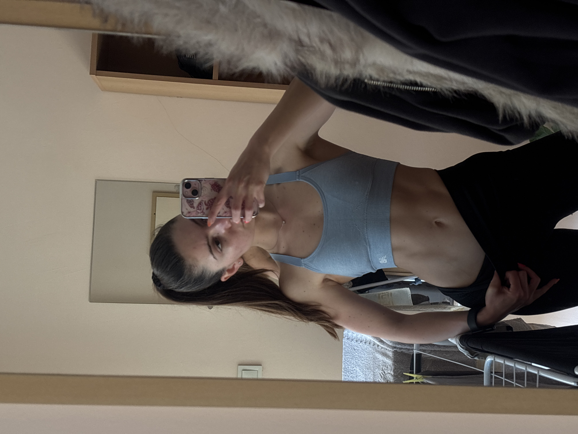 A person stands indoors, possibly in a bedroom, facing a mirror and holding a phone to take a selfie. The person is wearing a light blue sports bra and black leggings. The background includes a wardrobe with a shelf, a mirror, and a possible ironing board and clothes rack. The location is Unknown, Unknown.