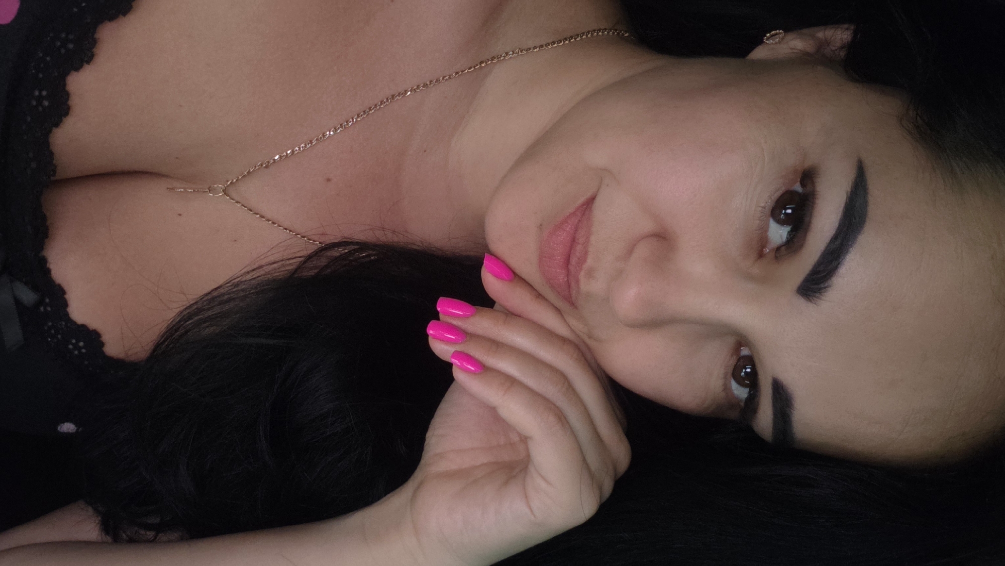 A close-up portrait of a woman with dark hair and brown eyes is shown. She is wearing a gold chain with a pendant and a black lace bra. Her hand, with bright pink nail polish, rests on her chin. The background is dark. The image location is Unknown, Unknown.