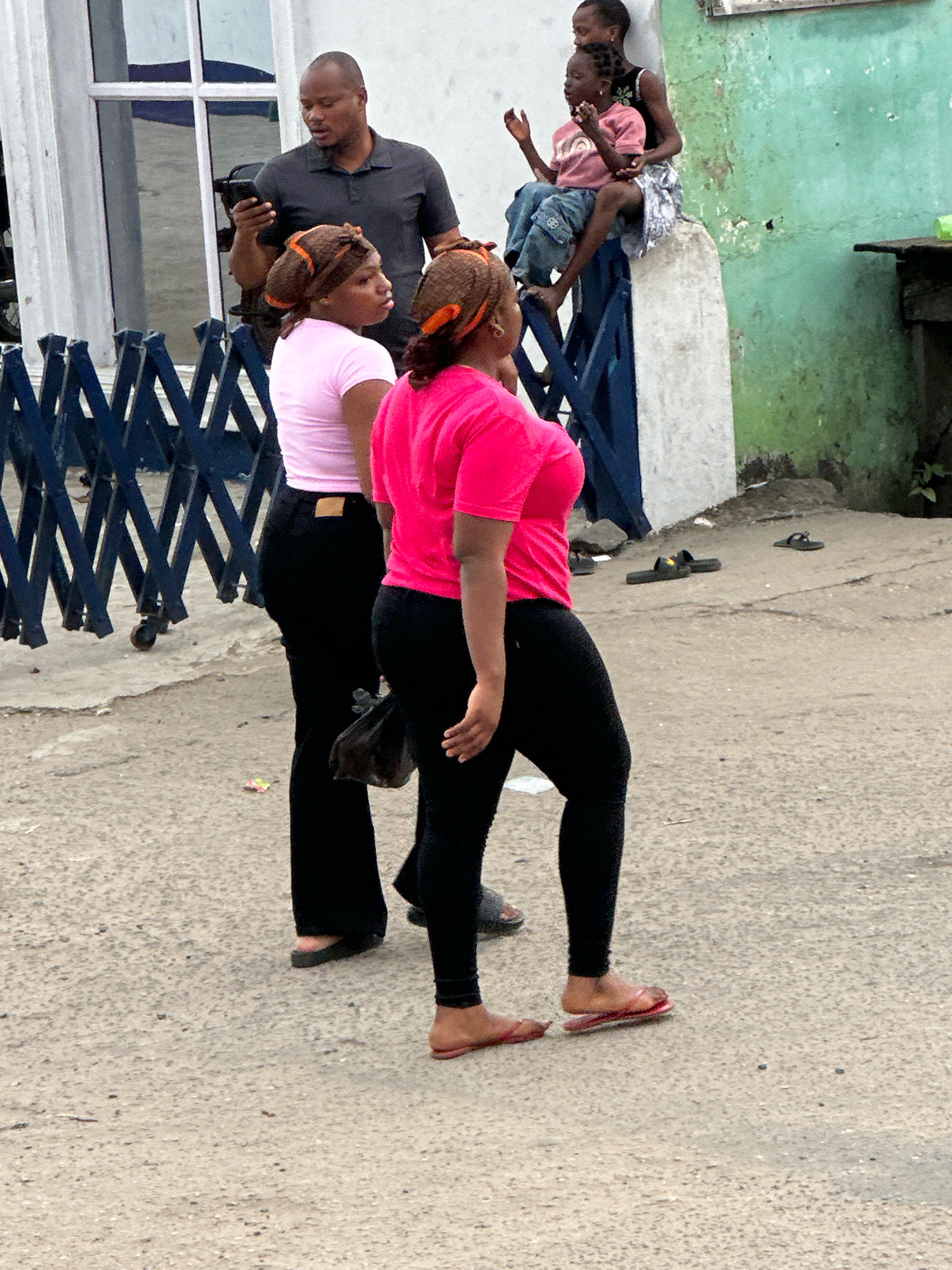 In Ajara, Nigeria, a man in a dark grey polo shirt holds a black smartphone in his left hand and looks down at it. Two women in black leggings and brightly colored tops stand facing away from the camera. The woman on the left wears a pink t-shirt, and the woman on the right wears a bright pink t-shirt. Both women have their hair styled with headscarves. In the background, two young children, one in a pink shirt and the other in a black tank top, sit on a ledge. The environment is an outdoor, urban setting with a concrete ground and a building with a distressed green painted wall. A metal accordion gate is visible on the left side of the image.