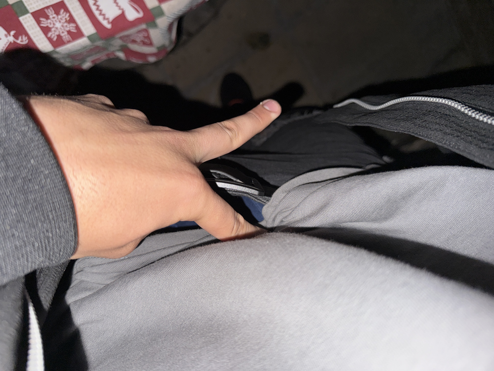 The image provides a close-up, top-down perspective of a human right hand, exhibiting a light-to-medium skin tone, positioned centrally. The fingers are slightly splayed, gripping or interacting with a dark, reflective object, consistent with a mobile phone, as it is placed into or retrieved from a pocket or compartment. This scene is situated in Varna, Bulgaria.
The hand is framed by multiple textile elements. A dark grey ribbed fabric, likely a sleeve, is visible in the upper left. A significant portion of the left and lower mid-section is covered by a light grey, soft, and somewhat rumpled fabric, consistent with casual trousers or sweatpants. To the right and beneath the hand, a black textured fabric is present, featuring a distinct light-colored, braided cord or zipper detail running vertically, indicative of a garment pocket or a bag. A small area of dark blue fabric is also visible beneath the fingers.
In the upper right background, a patterned textile, possibly a blanket or tablecloth, displays a red, white, and green checkered design adorned with white holiday motifs, including reindeer and snowflakes. The lower right background is dark and out of focus, revealing a blurred reddish object, potentially a shoe, resting on a dark surface. The environment is dimly lit, casting soft shadows, suggesting an indoor setting.