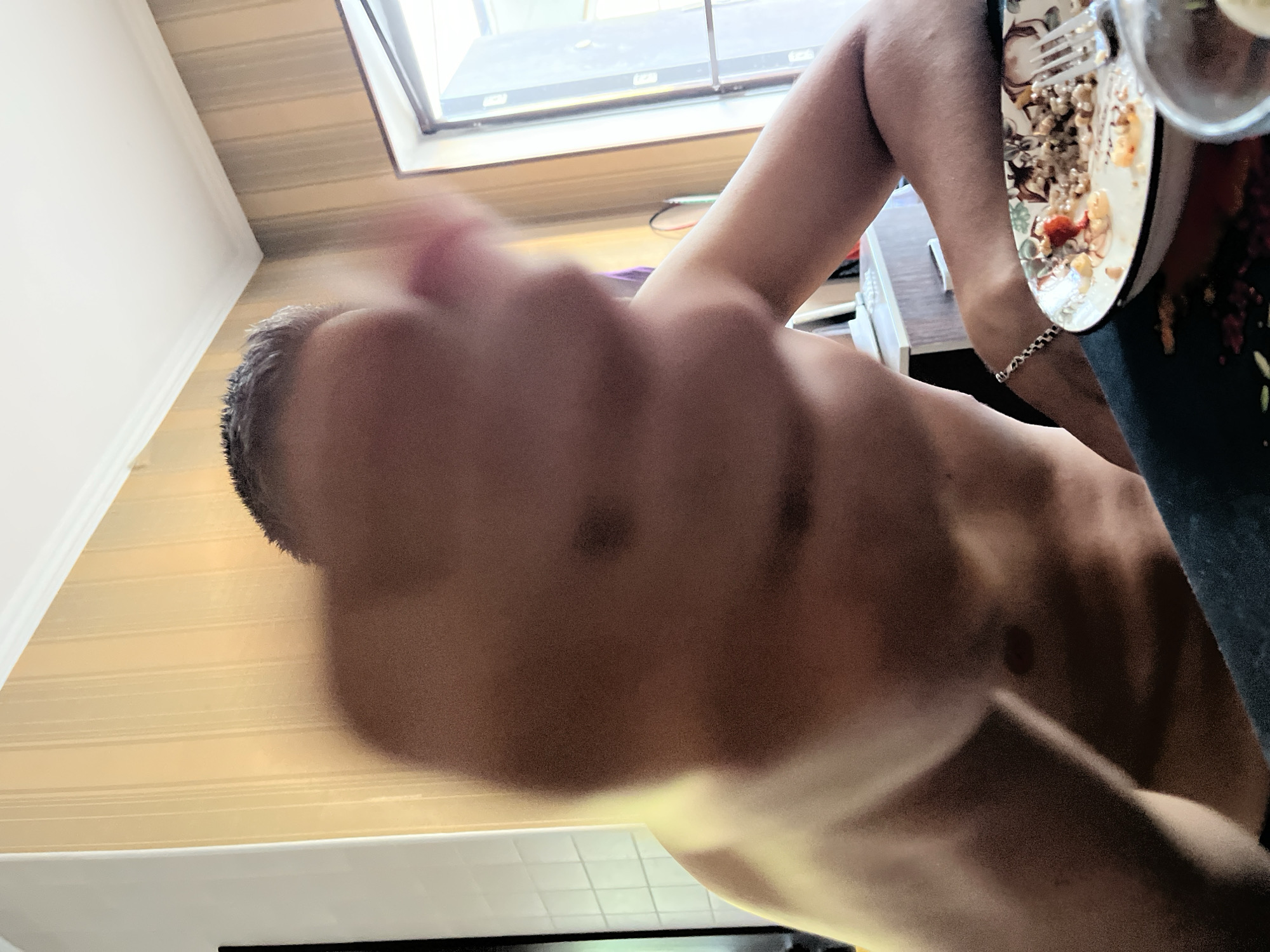 The image depicts a close-up, out-of-focus view of a shirtless person's chest and arm, with their hand reaching towards the camera. The person appears to be seated at a table with a plate of food and a fork. The background shows a wall with vertical striped wallpaper and a window. There is no visible indication of the city of Shymkent, Kazakhstan, or any specific urban elements. The lighting is warm, suggesting indoor illumination or natural light from the window.
