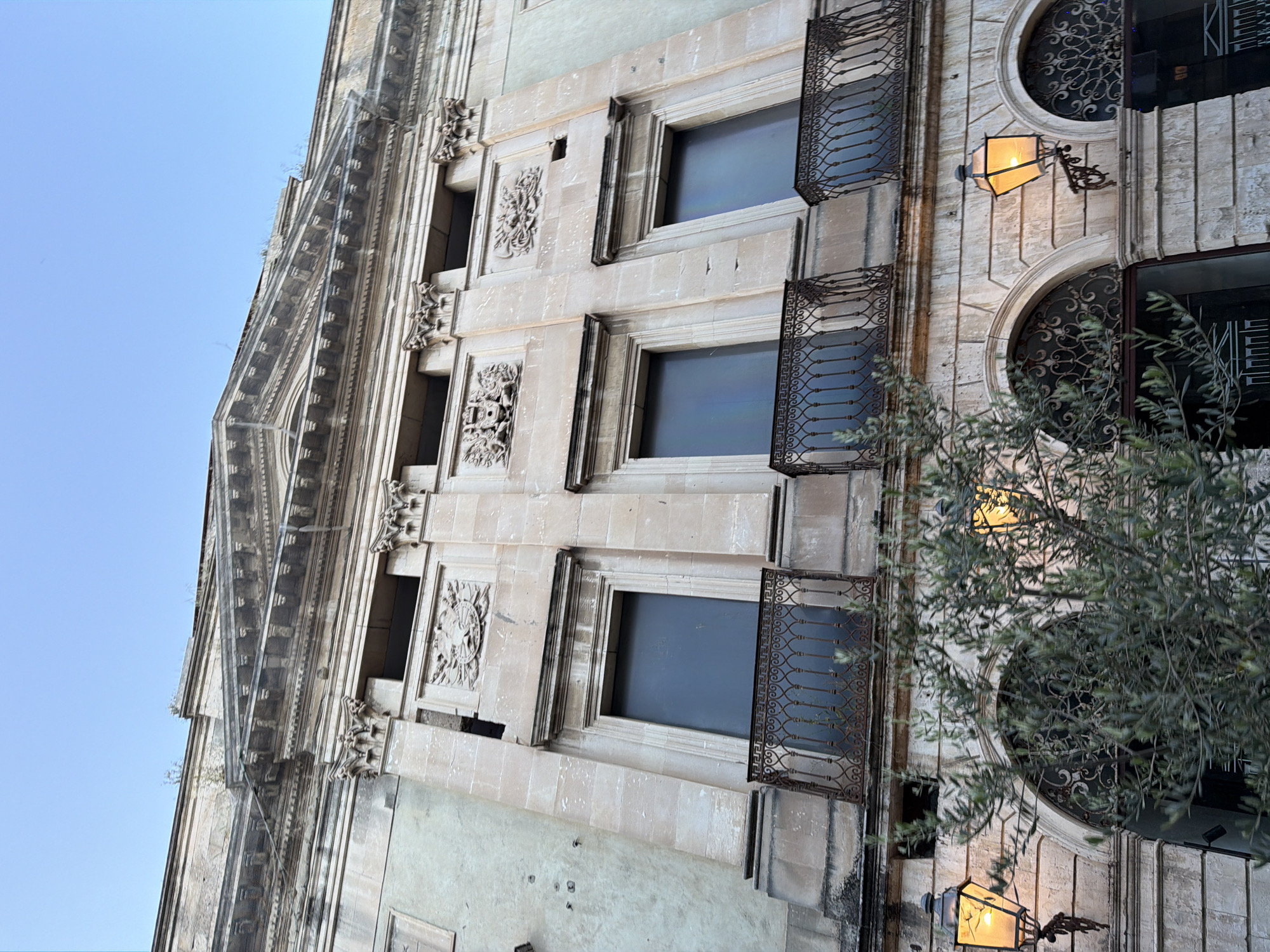 The image shows a section of a building in Acireale, Italy. The facade is made of light-colored stone with ornate details, including decorative carvings around the windows. The windows are rectangular, except for the ones on the right, which are arched. Each window has dark glass and a wrought-iron balcony. A street lamp is mounted on the building between the arched windows. A tree with dense foliage is partially obscuring the lower right of the image. The sky is visible in the upper left corner.