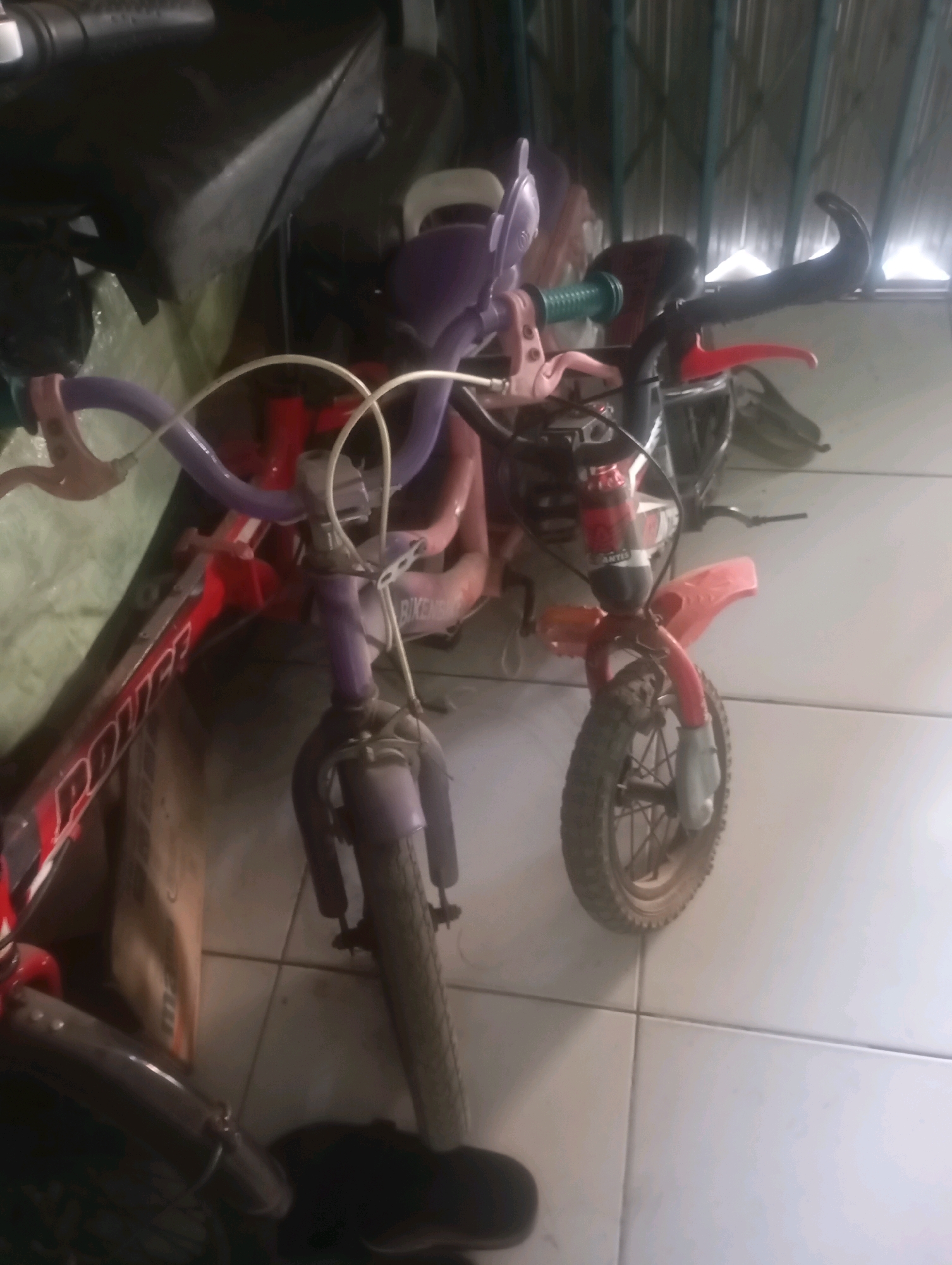 In Babelan, Indonesia, two children's bicycles are leaning against a wall in an indoor space with a tiled floor and a corrugated metal ceiling. The bicycle on the left is red and has "POLICE" written on its frame. It has purple handlebars with white grips and a purple seat. The bicycle on the right is red and appears to be a dirt bike or motocross style bicycle, with a small red fender over the front wheel and a black fender over the rear wheel (mostly obscured). It has purple handlebars and a purple frame visible. Both bicycles appear to be used and are positioned in a cluttered environment, with other objects, possibly bags or tarps, visible in the background.