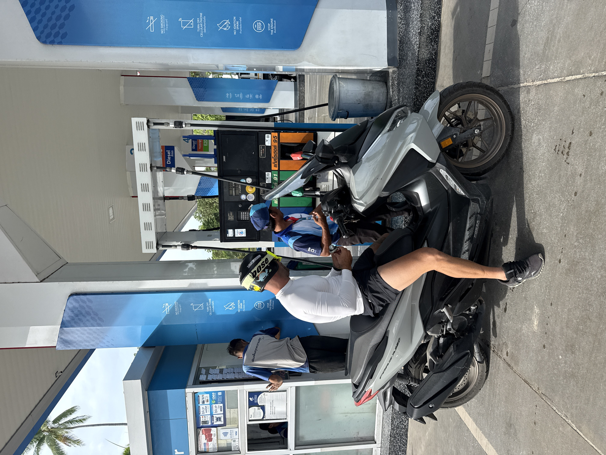 The image depicts a scene at a gas station in the Ko Samui District of Thailand. A man wearing a white long-sleeved shirt, black shorts, a helmet, and athletic shoes is seated on a gray scooter. He is holding a smartphone in his hands and appears to be looking at it. In the background, two men in blue and white uniforms are attending to a gas pump. One man is standing near the pump with his back to the camera, while the other, wearing a blue cap, is facing the scooter. A gas pump with various fuel options, including "Diesel" and "95," is visible. Several safety signs are displayed on a blue column, including "NO SMOKING," "TURN OFF CELLULAR PHONE," "NO IGNITION SPARK," and "STOP THE ENGINE." The environment appears to be an outdoor covered area of the gas station, with a palm tree visible in the upper left corner. The ground is paved with concrete.