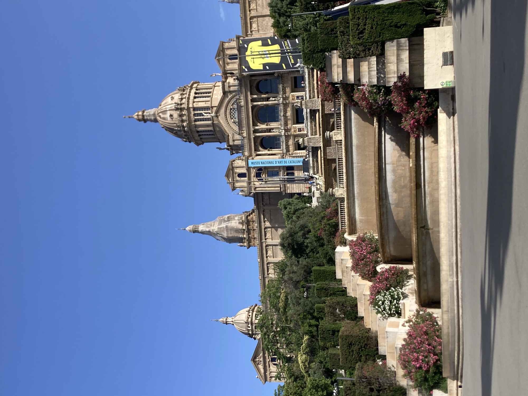 The image depicts the Palau Nacional in Barcelona, Spain, a grand building housing the National Art Museum of Catalonia. The building is a prominent architectural landmark with multiple domes and classical facade details. In front of the building, a multi-tiered fountain cascades down a series of stone steps, adorned with lush greenery and flowering bushes in shades of pink and white. The scene is bathed in bright sunlight under a clear blue sky, suggesting a pleasant day. The steps of the fountain are made of stone, and the water creates reflective surfaces. The building itself is made of stone and features large arched windows. On the facade of the building, two banners are visible; one is blue with text, and the other is yellow with a graphic of a t-shirt and text. There are some people visible on the upper terraces of the building, small figures against the vast structure. The foreground shows a paved area, possibly a plaza or walkway, with shadows cast by an unseen object. The overall impression is one of grandeur, nature, and a public space designed for enjoyment and cultural appreciation.