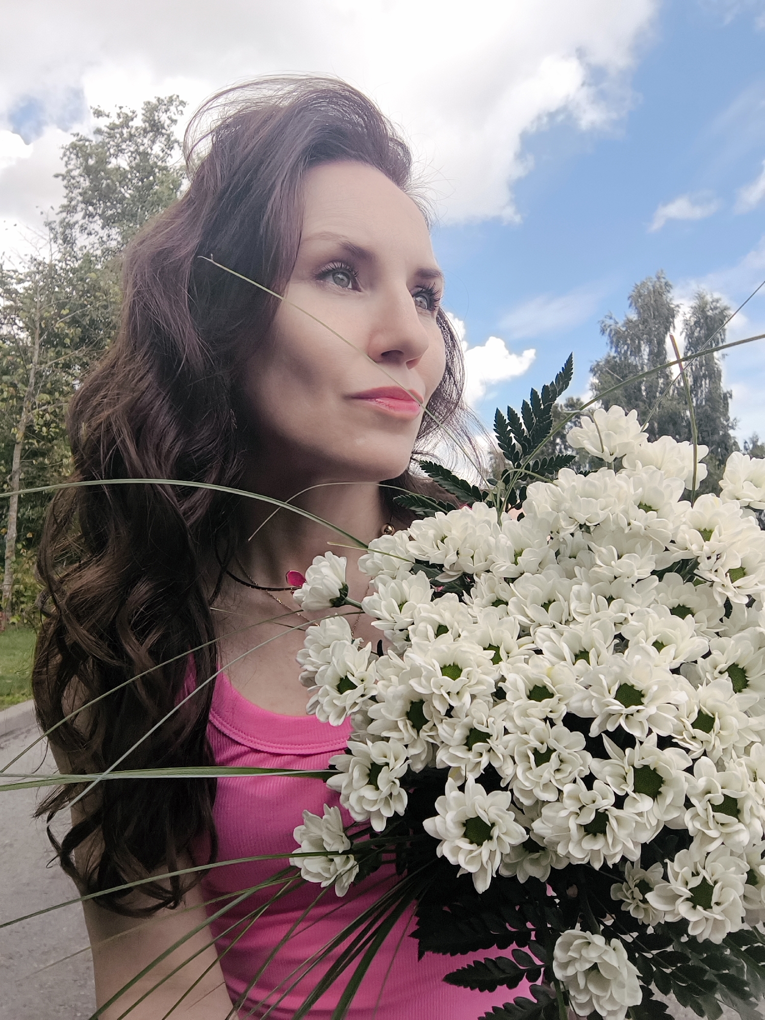 A medium shot captures a woman holding a bouquet of white chrysanthemums outdoors. She is positioned in the foreground, her profile facing right. Her long, dark, wavy hair cascades over her left shoulder. She wears a bright pink tank top. The bouquet of flowers is prominently displayed to her right, obscuring a portion of her body. In the background, trees and a bright blue sky with scattered clouds are visible. The ground beneath her appears to be a paved surface, possibly a sidewalk or road. The scene is illuminated by natural daylight.