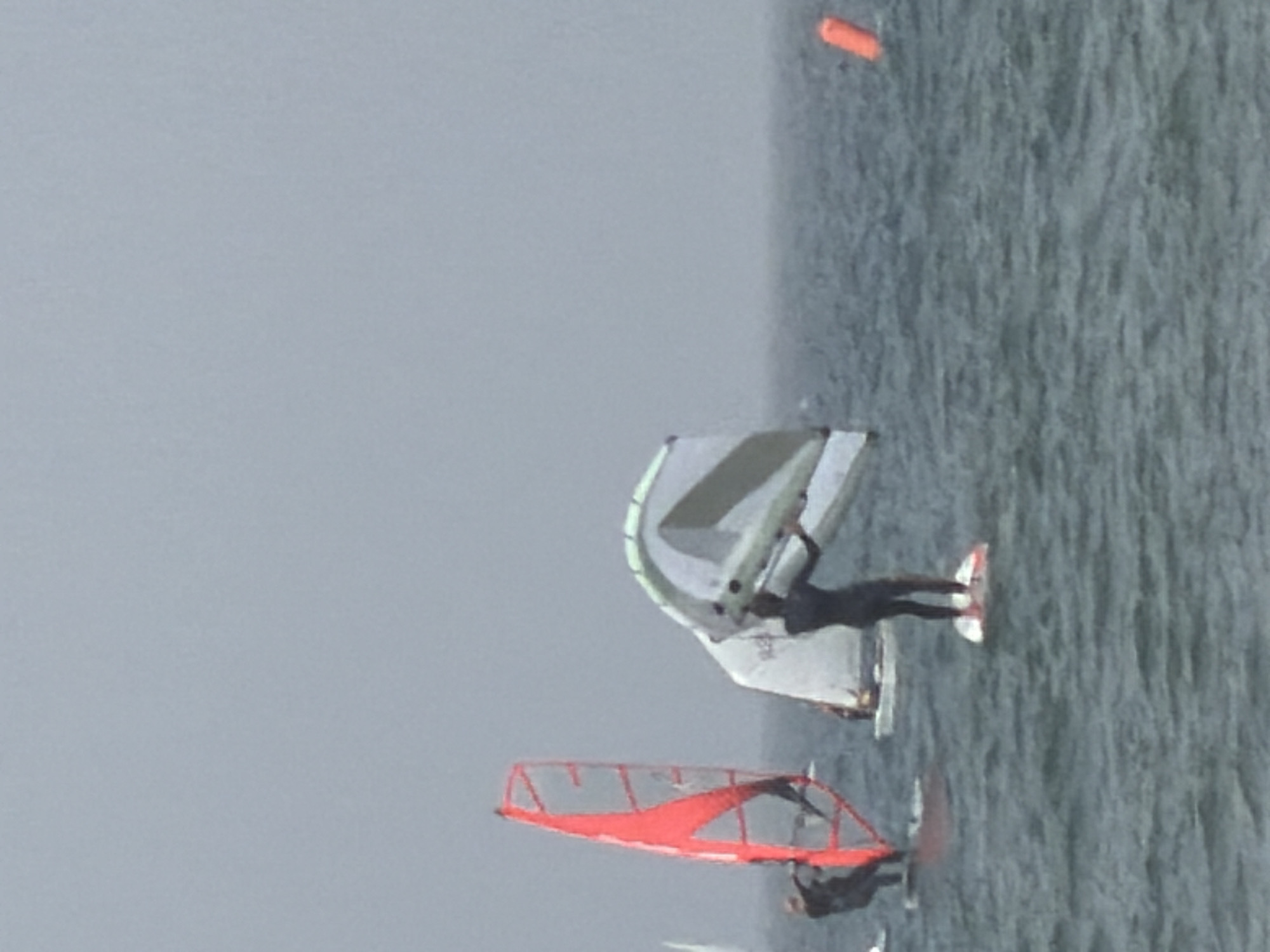 The image depicts two windsurfers on a body of water, possibly the Mediterranean Sea off the coast of Tel Aviv. The sky is overcast, creating a gray backdrop. One windsurfer has a red sail, and the other has a white and grey sail. An orange buoy is visible in the upper right corner. The sea is choppy.