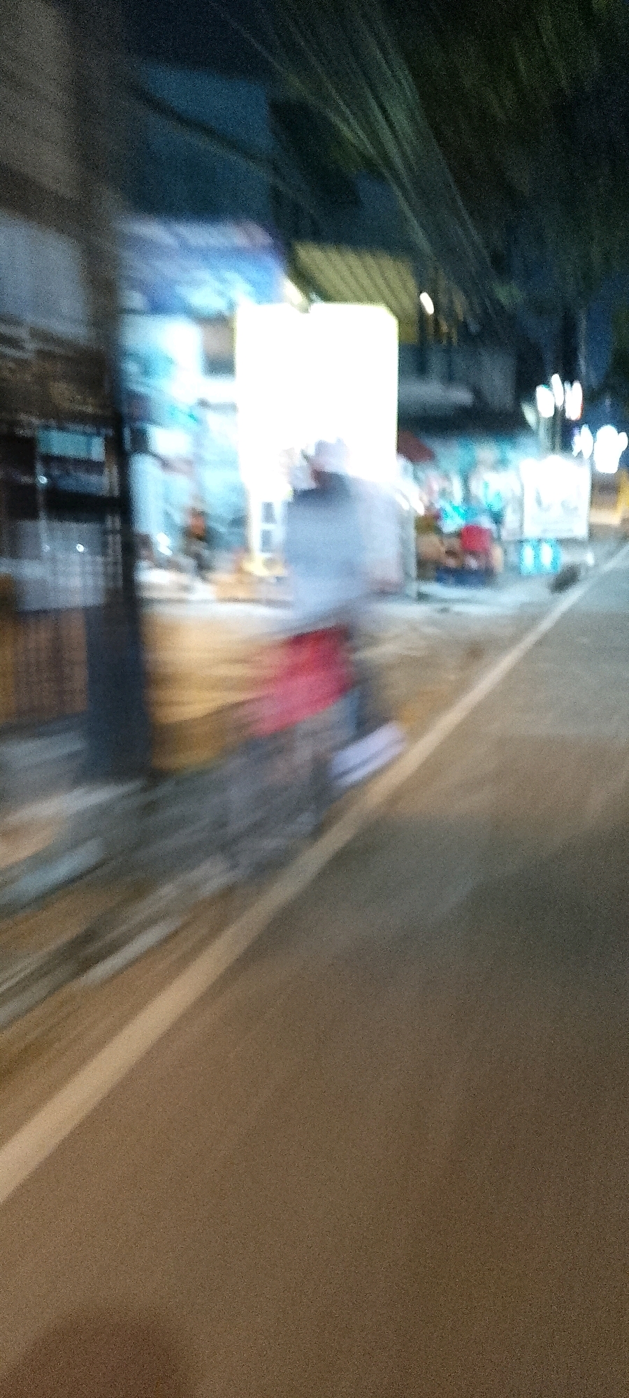The image depicts a blurry, nighttime scene of a street in Bekasi, Indonesia, likely a commercial or residential area with shops and possibly food stalls. The foreground is dominated by a street with a white dividing line. The background shows the blurred outlines of buildings, bright lights, and what appears to be trees or foliage overhead. There is a noticeable motion blur, suggesting the image was taken from a moving vehicle. A person or persons are visible as indistinct, blurred figures, one with a red garment, in front of the shops. The overall impression is one of a busy, active street at night, with the blur emphasizing movement and the low light conditions.