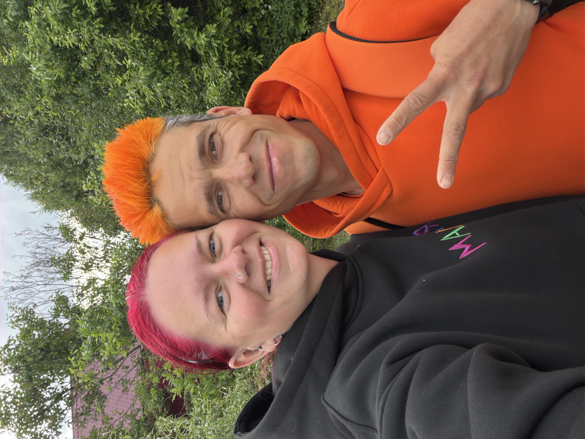 A vibrant selfie, rotated 90 degrees clockwise, captures two smiling individuals outdoors. The man, positioned in the upper portion of the frame (when viewed in its current orientation), has bright orange hair with visible grey roots and wears an orange hoodie. He smiles warmly at the camera and makes a peace sign with his right hand, which is visible towards the right side of his body. His eyes are dark brown.
Next to him, in the lower portion of the frame, is a woman with striking bright pink hair. She has a wide, cheerful smile, revealing her teeth, and sports a silver nose ring along with multiple silver earrings. She is dressed in a black hoodie. On the left side of her chest, colorful embroidery spells out "MAM" followed by an indistinct character, using shades of purple, green, and orange/yellow.
The background consists of lush green foliage from trees and bushes, indicating an outdoor setting, likely a park or garden in Chaikovsky, Russia. A cloudy sky is visible through the leaves in the upper left corner. Both subjects convey joy and closeness through their happy expressions, suggesting a cheerful moment captured on a bright day.