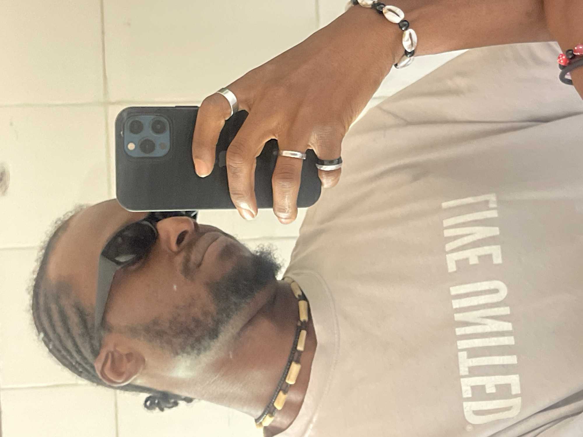 The image is a selfie taken by a Black male in what appears to be a bathroom. He is wearing a light-colored t-shirt with the words "LIVE UNITED" printed in reverse and in capital letters. He has his hair braided and is wearing dark sunglasses. He is also wearing a beaded necklace and several rings on his fingers. The background shows a tiled wall, typical of a bathroom. There is no indication of the specific location in Jos, Nigeria.