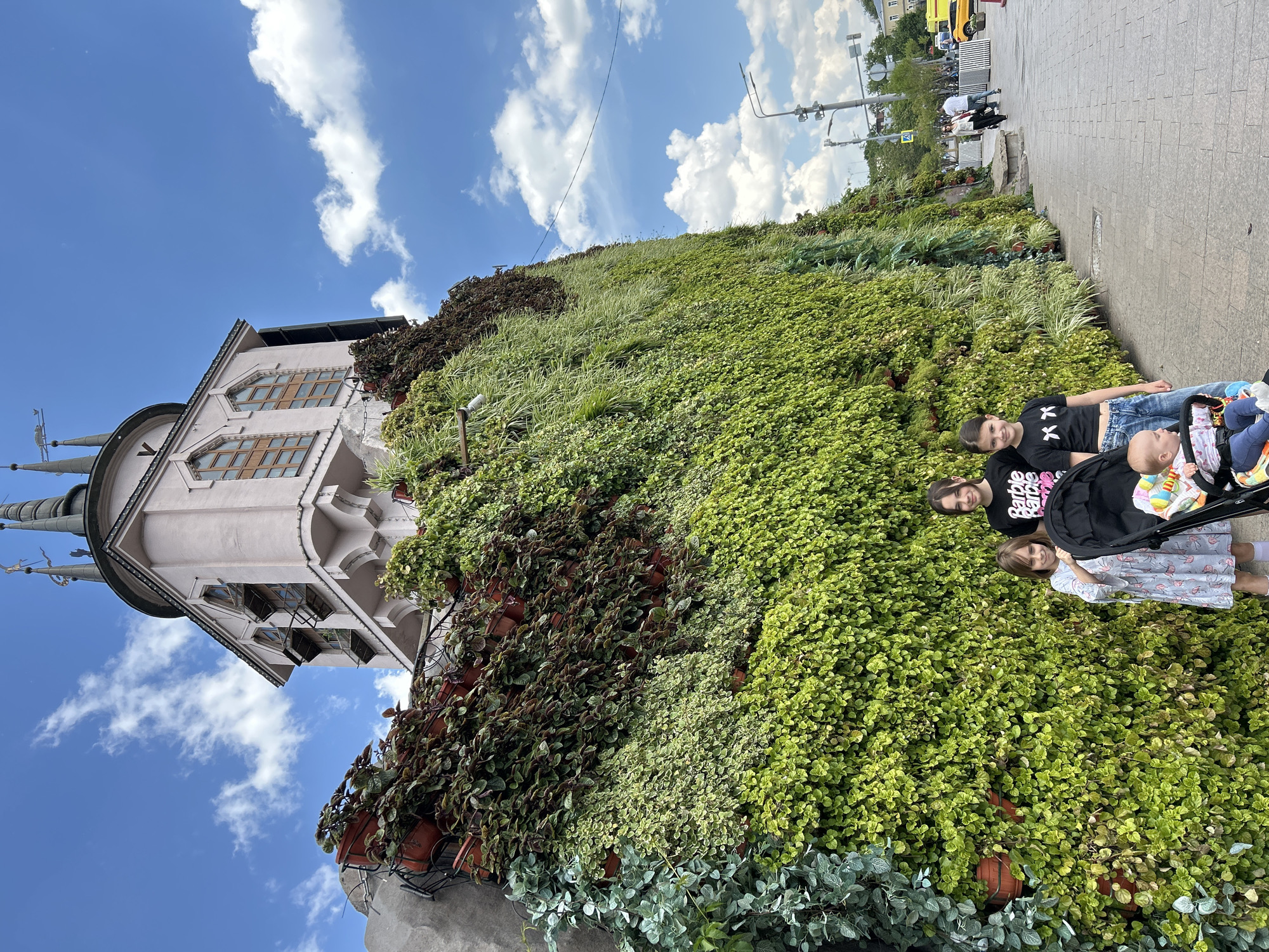 The image displays an urban outdoor scene featuring a distinctive building, a large living green wall, and several people on a sunny day in what is identified as Moscow, Russia.
In the foreground, on a paved sidewalk, are four individuals. An adult or older teenager, wearing a black t-shirt with two white "xx" symbols and blue jeans, smiles towards the camera. Beside her, another individual in a black t-shirt featuring the "Barbie Barbie" logo also smiles, pushing a black stroller containing a baby. A young girl, dressed in a light-colored, possibly flamingo-patterned dress, stands near the stroller, also smiling towards the viewer. The baby in the stroller is partially visible, wearing colorful clothing.
To the left, a unique light pink or beige building with intricate architecture stands prominently. It features ornate hexagonal windows and multiple spires or towers, contributing to its distinctive appearance.
A dominant feature of the scene is the expansive living green wall that covers a significant portion of the image. This wall is densely populated with a variety of plants, including vibrant green groundcover, taller grasses, and darker leafy foliage. Some plants are visibly housed in terracotta pots integrated into the wall's structure, creating a dynamic and lush facade.
The sky above is a clear blue with scattered white clouds, indicating bright, sunny weather during the daytime. In the background, on the street and sidewalk beyond the green wall, other people, streetlights, traffic signals, and a yellow vehicle (likely a taxi) are visible, reinforcing the bustling urban setting. The facial expressions of the foreground subjects convey a pleasant and cheerful atmosphere.