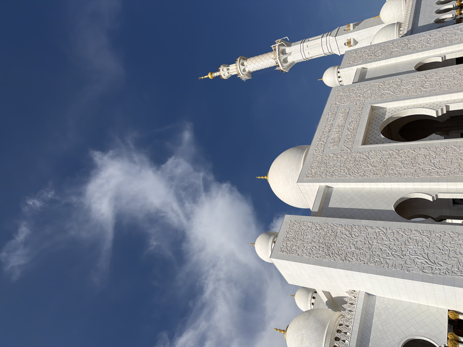 The image presents a striking, low-angle view of a grand, white architectural structure, likely a mosque, set against a vibrant blue sky with scattered white clouds. The structure, predominantly white marble, is characterized by intricate decorative carvings and multiple domes with golden finials.
In the upper central to upper right portion of the image, a tall, slender minaret rises diagonally upwards, also crafted from white marble and featuring ornate details and a golden finial at its apex. Below the minaret and extending towards the bottom and right edges of the frame, the main body of the building is visible. It features large, smooth white domes, each topped with a golden spire. The walls of the building are adorned with highly detailed, floral-inspired carvings in relief, creating a rich texture. Arched openings and recessed sections are also visible, adding depth to the architecture.
The perspective is upward-looking, emphasizing the colossal scale and intricate craftsmanship of the building. The deep blue sky occupies the majority of the left and upper parts of the image, punctuated by wispy, white clouds that appear to float behind the structure. The bright natural light suggests it is daytime, casting clear visibility on the details of the white marble and the golden accents. No people or text are visible in the image.
