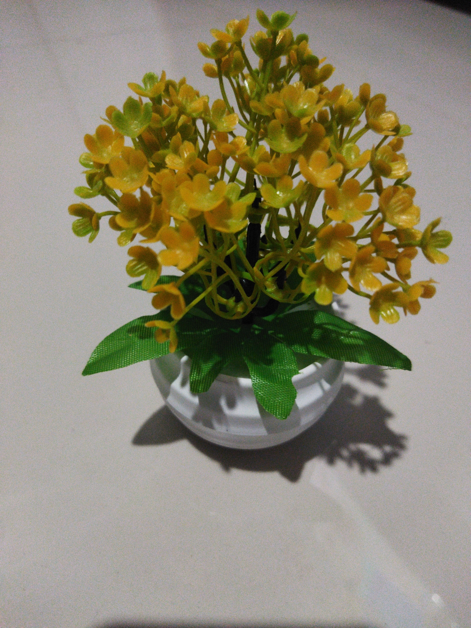 A close-up, high-angle shot shows a small, artificial potted plant with yellow flowers and green leaves. The plant is in a white, rounded pot, and the leaves are a bright, textured green. A shadow of the plant is cast on the light-colored, textured surface it sits on. The location is not visible or determinable from the image.