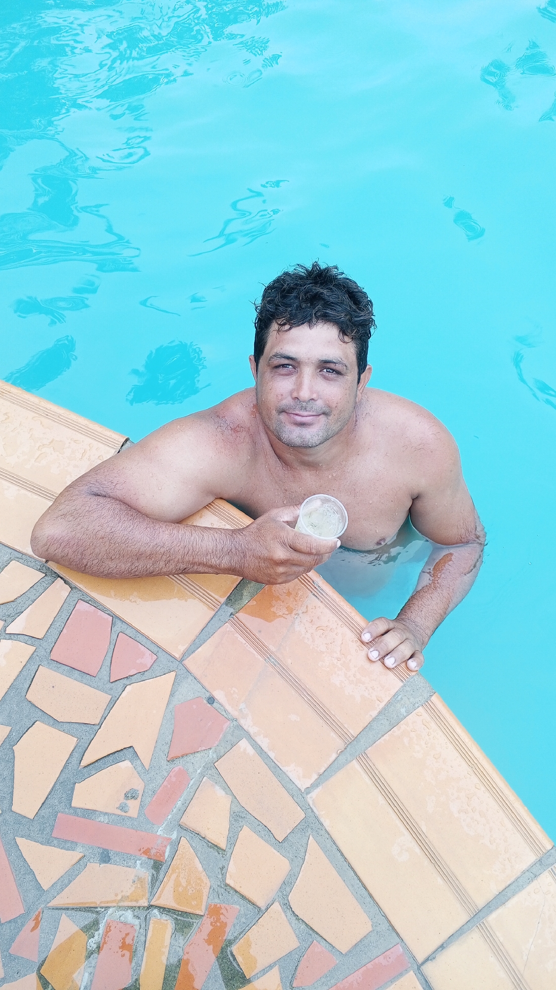 A man is in a swimming pool, holding a glass. The pool is lined with orange and beige tiles. The location is Unknown, Unknown.
