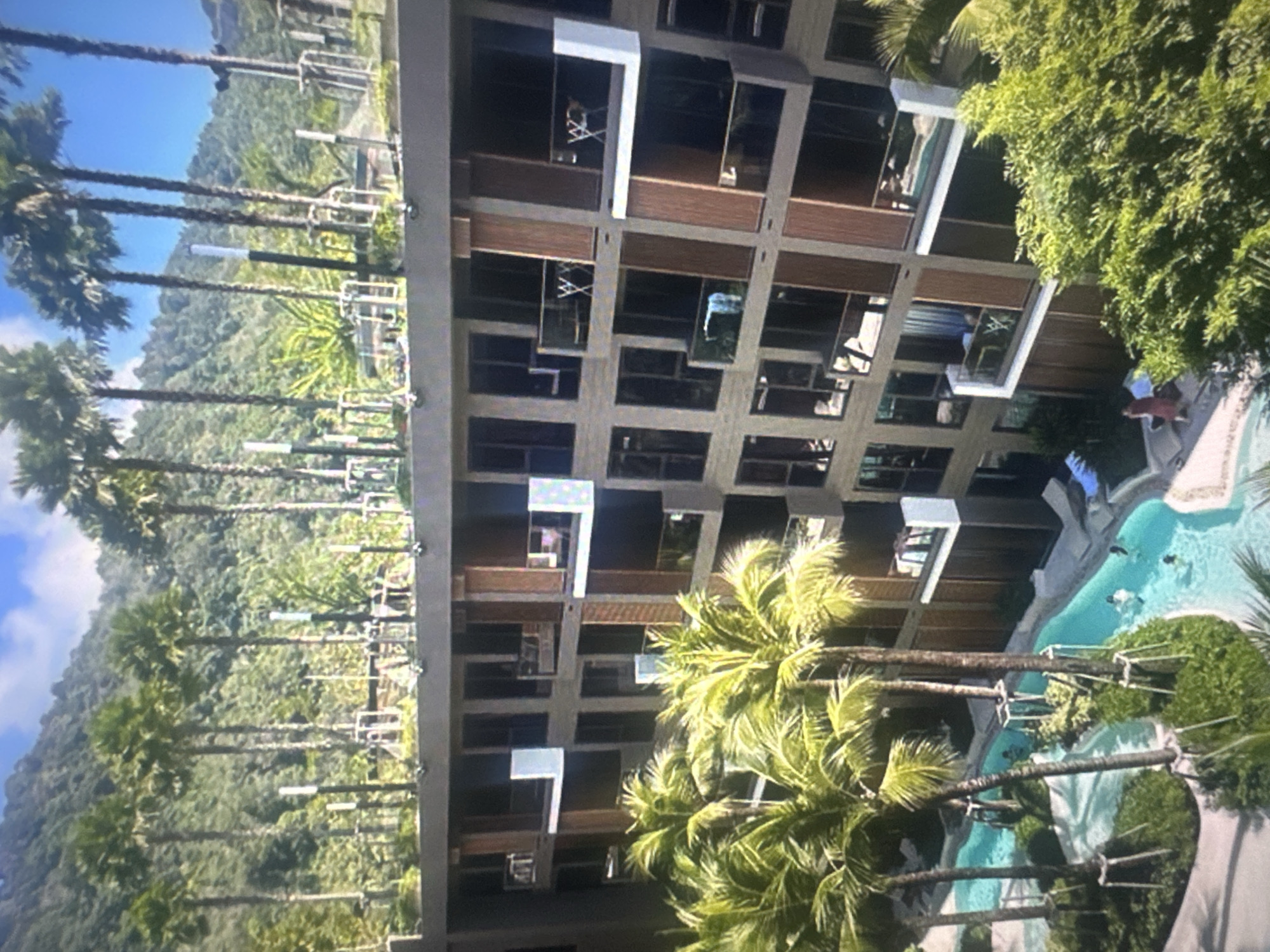 An aerial view captures a modern, multi-story resort building set against a backdrop of lush green mountains and a bright blue sky. Palm trees line the foreground, with their fronds fanning out over a winding, turquoise swimming pool. Several individuals are visible in the pool, enjoying the water. The building itself features a contemporary design with numerous balconies and large windows. On the rooftop of the building, a row of palm trees stands tall, and what appears to be outdoor exercise equipment is present. The overall scene depicts a luxurious tropical vacation destination.