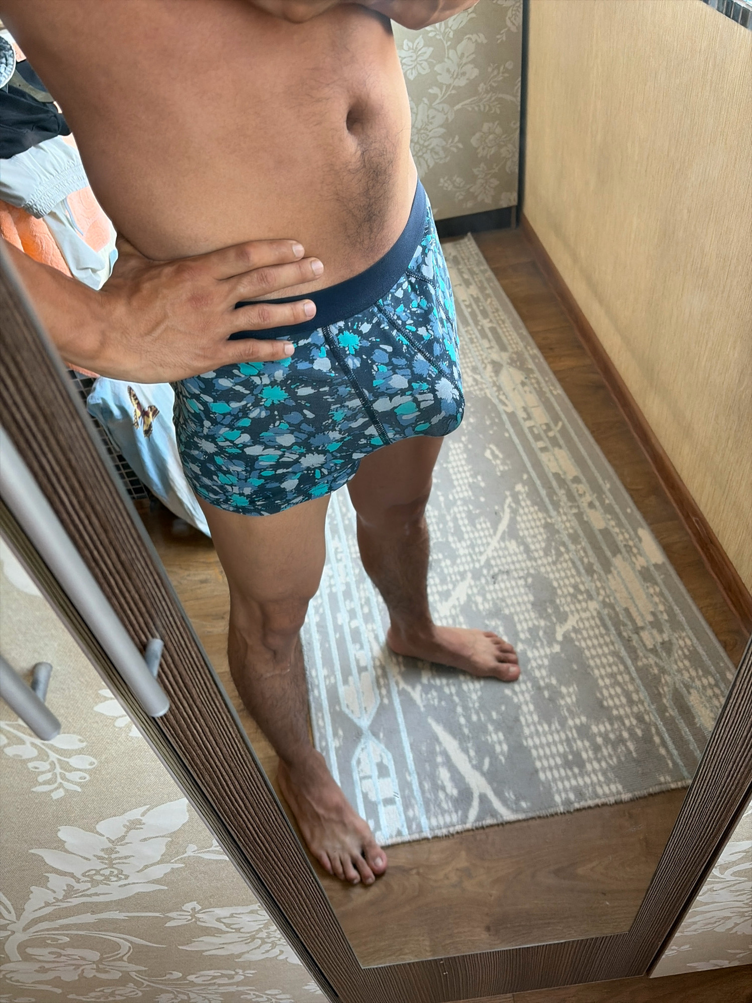 A person's torso and legs are visible in a reflection in a full-length mirror. The person is wearing patterned boxer briefs and is standing on a rug in a room. The room has wooden flooring and wallpaper with a floral pattern. In the background, a wardrobe is also reflected.