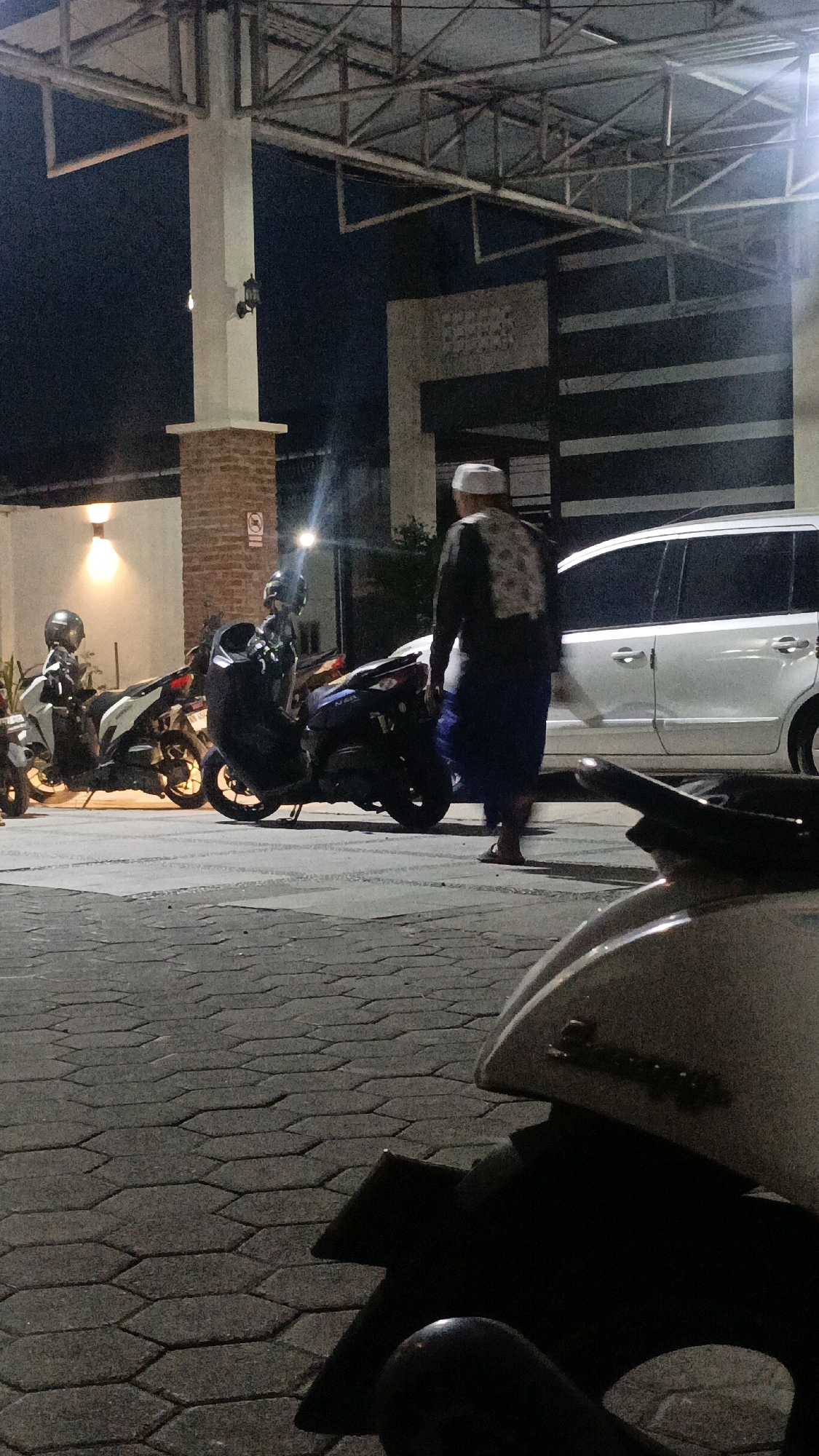 A man wearing a white skullcap and a dark patterned long-sleeved shirt over a blue sarong walks away from the camera in front of a white van. Several motorcycles are parked on a hexagonal paver stone surface. A modern building with a metal roof structure is visible in the background. The scene appears to be outdoors at night, with artificial lighting present.