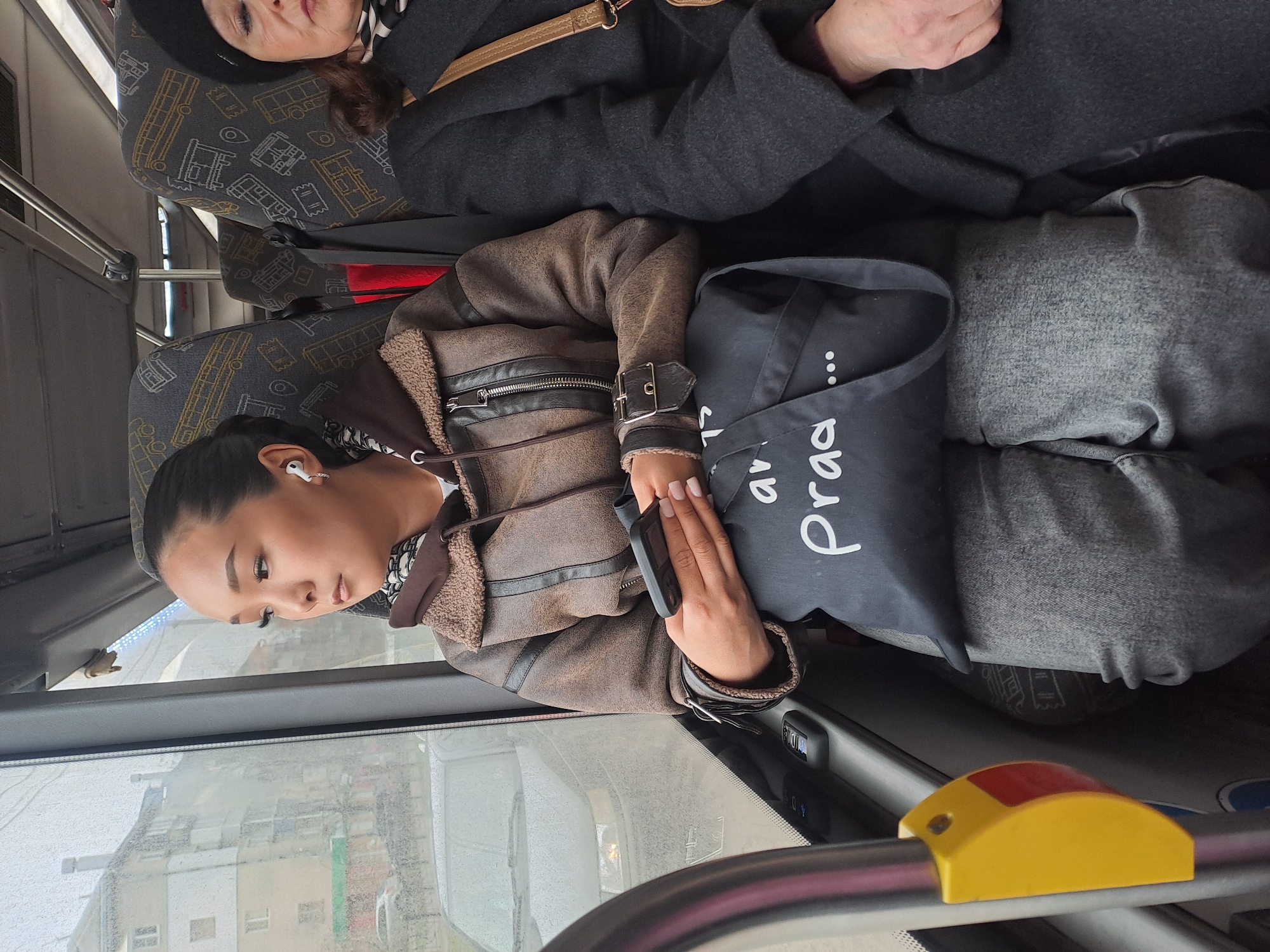 A young woman is sitting on a bus. She is looking out the window. She is wearing a brown jacket and gray pants. She has a black bag with "Praga" written on it. An older woman is sitting next to her wearing a black hat and gray coat. The bus has seats with a pattern of yellow and white. The bus window is partially obscured with condensation/water droplets. Outside the window, buildings are visible. The city is Elektrostal, Russia.