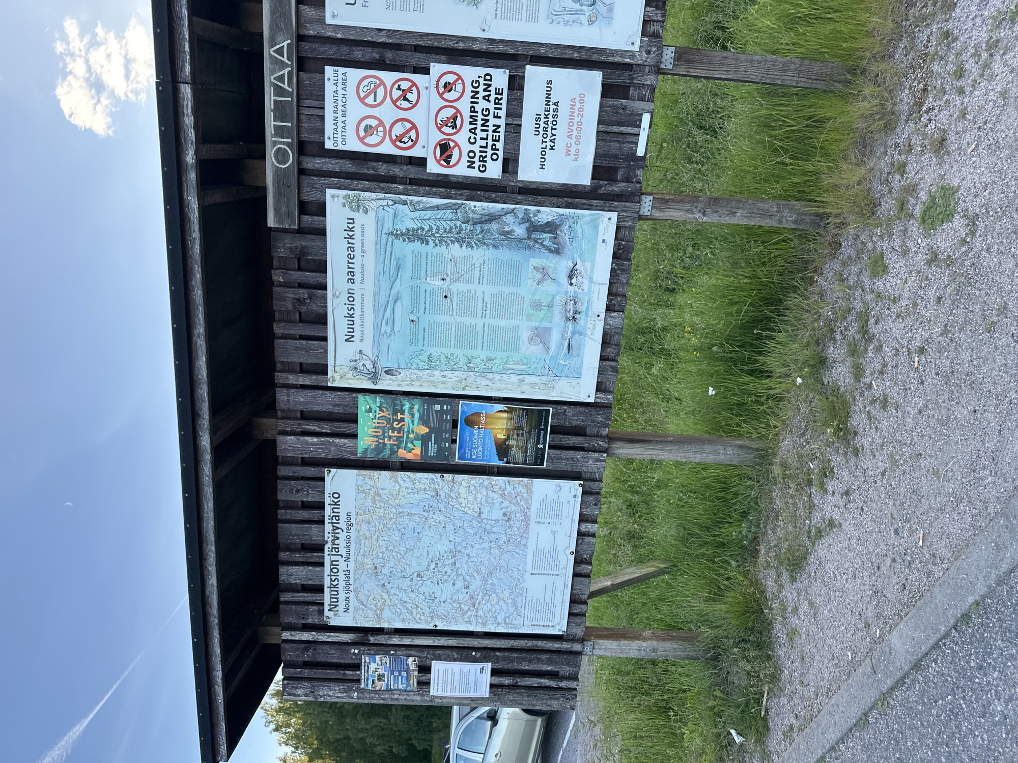 The image shows an outdoor information board at the Nuuksio National Park, located in the municipality of Espoo, Finland. The board is made of wood and displays several signs and maps, including a map of Nuuksio Lake Area, an information poster about "Nuuksio aarrekorkku" (Nuuksio treasure chest), and various cautionary signs regarding camping, grilling, and open fires. A sign in Finnish and English indicates "OITTAA BEACH AREA" and prohibits camping, grilling, and open fires. Another sign announces that the "UUSI HUOLTORAKENNUS" (new service building) is in use with WC opening hours from 06:00 to 20:00. The board is situated in a grassy area with trees in the background, and a parking lot with a car is visible on the left side of the image. The sky is clear with some clouds and a crescent moon is visible.