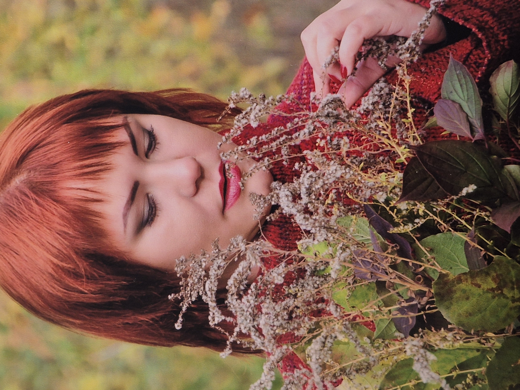 An outdoor, medium shot shows a woman with vibrant red hair and bangs, looking down at a bouquet of dried flowers and green and purple leaves she is holding. Her face is partially obscured by the foliage. She is wearing a textured red sweater. The background is blurred, suggesting an autumn setting with muted green and yellow tones. The woman's makeup includes dark eyeshadow and bright red lipstick. Her fingernails are painted a matching red. Her right hand is reaching into the bouquet. The image appears to be a portrait taken in Schwäbisch Gmünd, Germany.