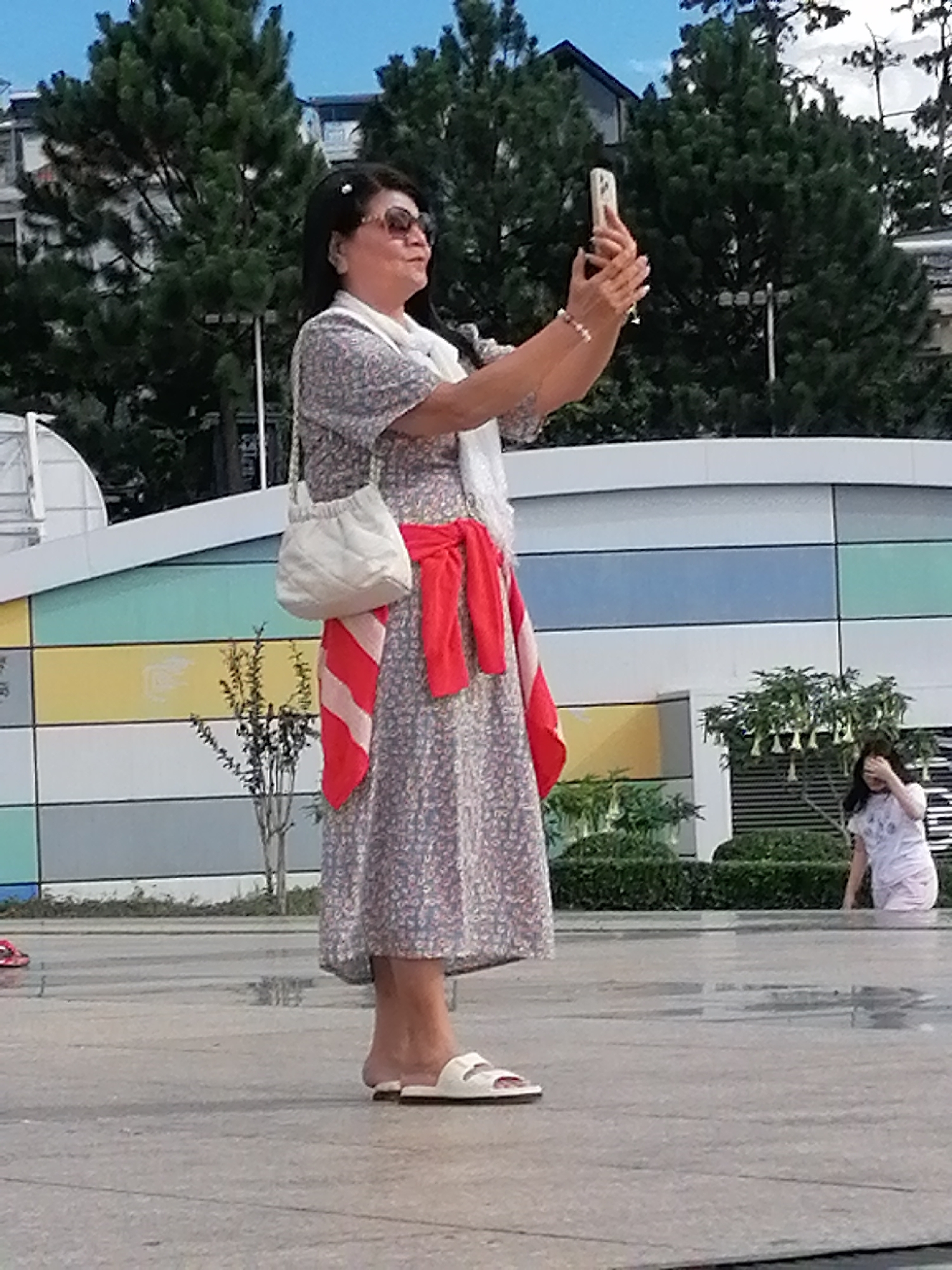 An East Asian woman is standing outdoors in what appears to be a public square in Da Lat, Vietnam, possibly near a fountain given the wet pavement. She is facing right, holding a white smartphone in her right hand, angled to capture a selfie. She wears a floral print dress, a red scarf tied around her waist, a white quilted crossbody bag, and white sandals. She has dark hair pulled back, adorned with a white clip, and is wearing sunglasses. In the background, on the right, a young girl in a light-colored t-shirt and shorts is partially visible, covering her eyes with her hand as she looks towards something out of frame. The environment includes a modern building with colorful horizontal panels and lush green trees.