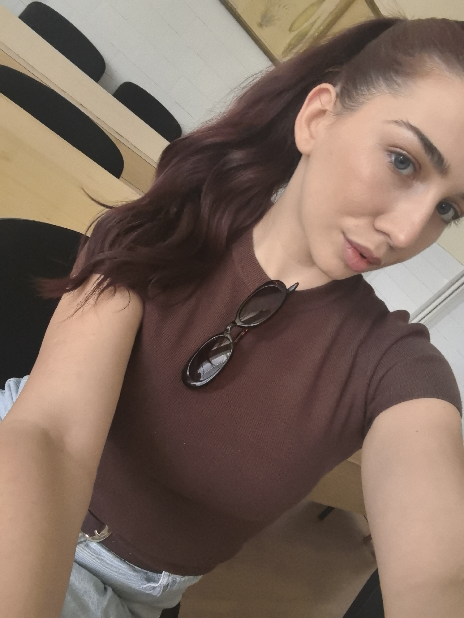 A woman with reddish-brown hair pulled back in a ponytail is taking a selfie. She is wearing a brown ribbed t-shirt and light blue jeans. A pair of dark sunglasses is resting on her t-shirt, over her left shoulder. Her left arm is bent and resting on her lap, while her right arm is extended to take the photo. The background appears to be an indoor setting with tiered seating, possibly a lecture hall or classroom. The lighting is natural, coming from the right side of the frame. No information about the city of Bogomilovo, Bulgaria, can be gleaned from the image.