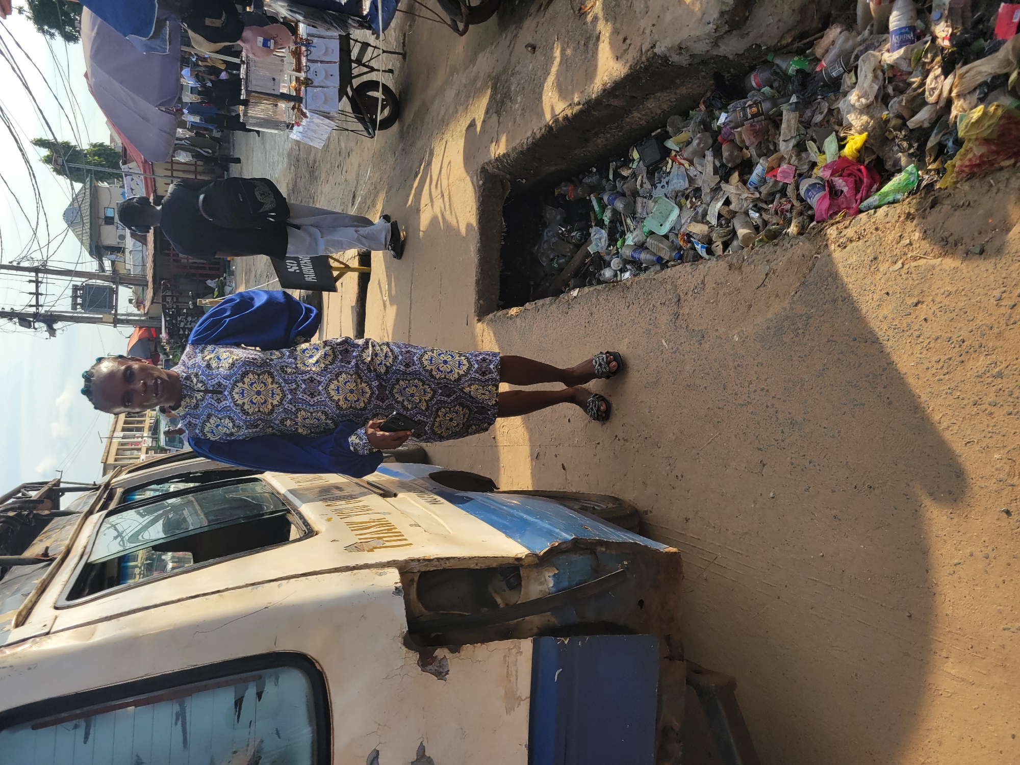 In the city of Egbagu, Nigeria, a woman stands on a dirt road beside a dilapidated white van. The van, with its rear damaged and showing exposed metal, has "THANK YOU GOD" painted in gold on its side. The woman wears a blue and gold patterned dress with voluminous sleeves and black sandals. To her right, a narrow trench is filled with a large amount of refuse, including plastic bottles, bags, and assorted trash. In the background, a man with a black backpack walks away from the camera, and market stalls with umbrellas and merchandise are visible, along with power lines and buildings. The lighting suggests it is daytime, with shadows cast on the ground.