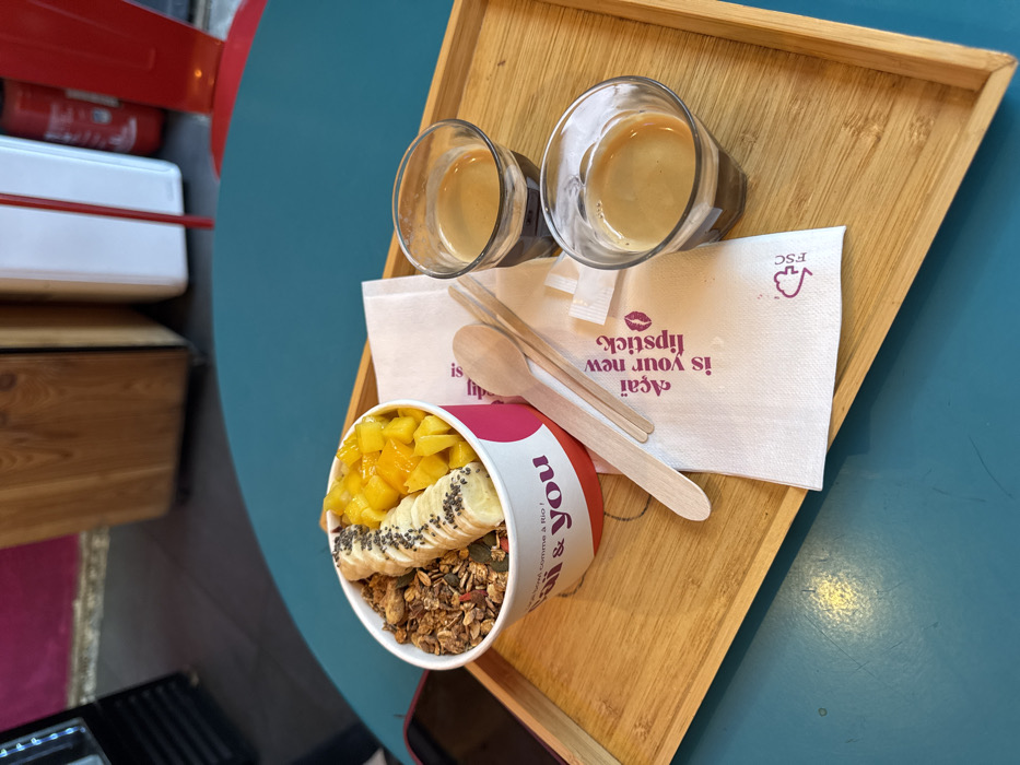 The image depicts a close-up, elevated view of a meal set on a teal-colored table within what appears to be an Açaí & You cafe in Paris, France. A light-colored wooden serving tray occupies the center, holding a white paper bowl, two small wooden utensils, a paper napkin, and two glasses of espresso.
The paper bowl, branded with "Açaí & you" and "le bowl comme à Rio !", contains an açaí bowl preparation. The contents include a layer of granola, precisely arranged sliced bananas topped with chia seeds, and diced mango. Beside the bowl, two thin wooden spoons or spatulas rest on a folded white paper napkin. The napkin displays pink text reading "Açaí is your new lipstick" alongside a lipstick kiss mark graphic and an FSC certification logo.
Two clear, small glasses, each containing a single shot of espresso with a layer of crema, are positioned on the right side of the wooden tray. A dark-colored mobile phone is partially visible on the teal table surface to the left of the tray.
The background, largely out of focus, reveals elements of the cafe interior. These include a dark floor, a wooden cabinet or counter, a white vertical structure resembling a refrigerator, a portion of a red metal chair, and a red fire extinguisher mounted on a wall. No individuals are visible in the scene.