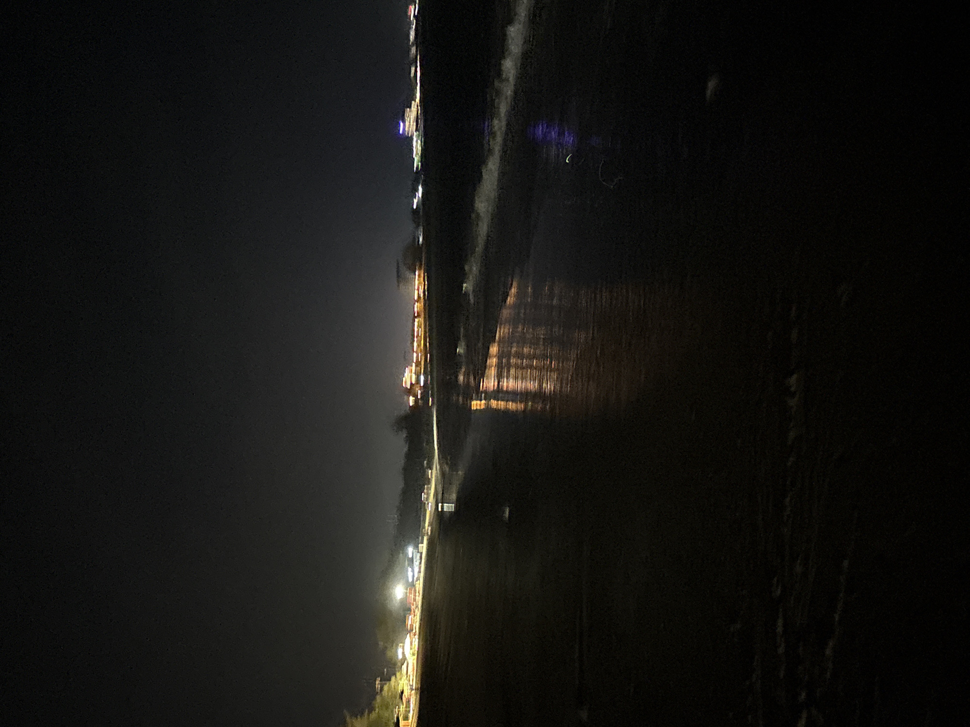 This nighttime image presents a panoramic view of a vast, dark body of water, likely a sea or a large bay, with a distant, illuminated shoreline. The perspective is from a dark, indistinct foreground, possibly a beach or landmass, looking out towards the water.
Along the distant left side of the frame, a long stretch of coastline is dotted with numerous artificial lights. These lights, appearing as bright points and streaks of warm yellow, orange, and some cooler white hues, suggest the presence of buildings, streetlights, or establishments lining the shore. These lights cast prominent, elongated reflections across the calm surface of the water, creating shimmering vertical lines that stretch into the mid-ground.
The water itself dominates the right half and much of the foreground, appearing mostly dark and reflective. Its surface seems relatively calm, with the primary visible features being the reflections of the distant lights. The immediate foreground is exceptionally dark, offering little discernible detail but implying a solid, dark ground leading to the water's edge.
The scene conveys a tranquil and expansive nighttime atmosphere. There are no identifiable people, animals, or specific activities taking place. The time of day is clearly night, with the entire scene illuminated solely by artificial light sources. No text is visible in the image. The location is contextualized as Dong Hai Ward, Vietnam, consistent with a coastal or waterfront setting.