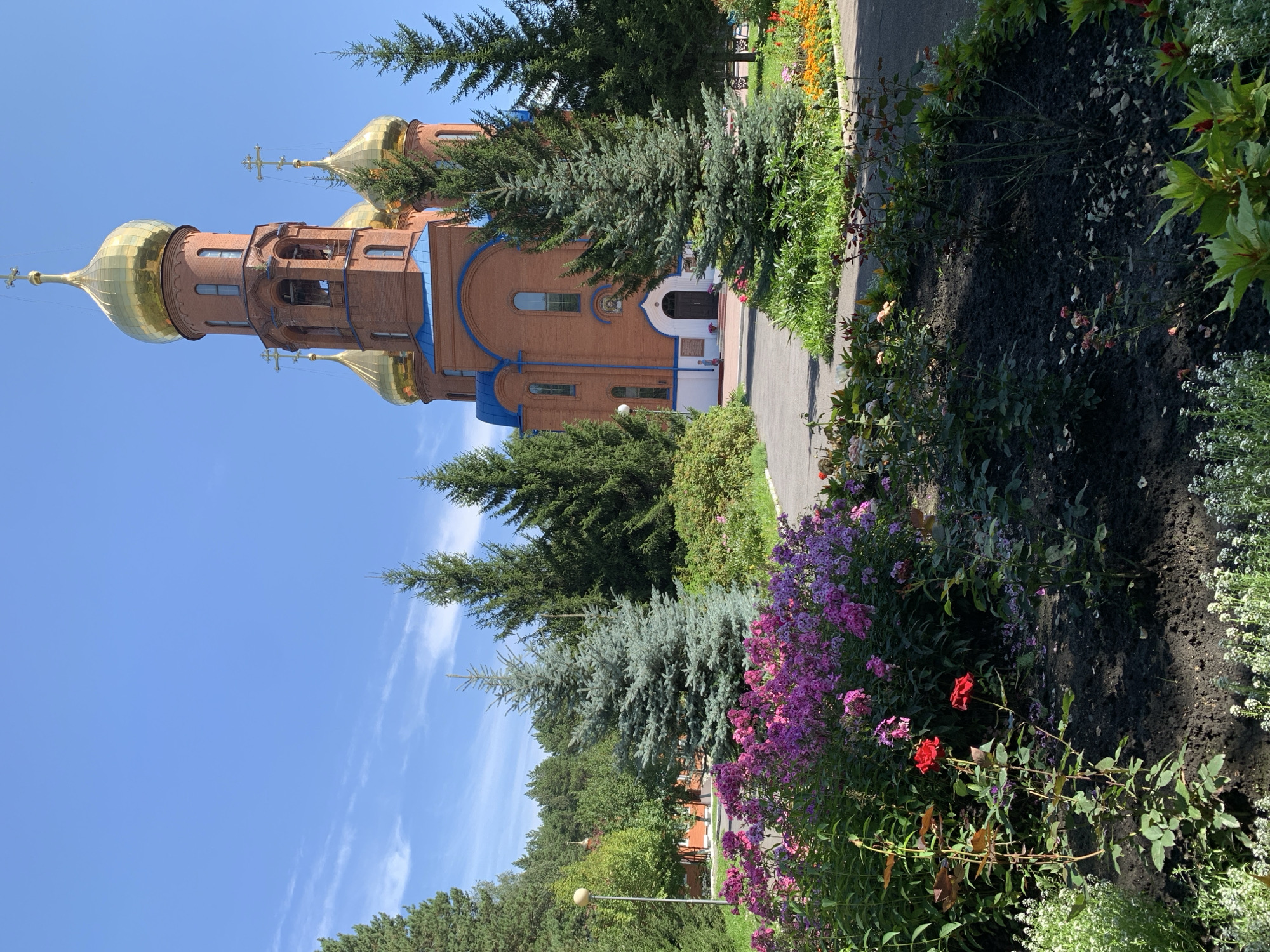 The image depicts the Church of the Nativity of the Most Holy Mother of God in Prokopyevsk, Russia, on a clear, sunny day. The church, constructed of red brick, features a central bell tower and multiple golden domes topped with crosses. Blue trim accents the roofline. In the foreground, a well-maintained garden bursts with colorful flowers, including pink and purple phlox, red roses, and smaller white blossoms. Various evergreen trees, including what appears to be a blue spruce, are interspersed throughout the scene, framing the church and gardens. A paved pathway leads towards the church entrance. The environment is lush and green, suggesting a temperate climate. No people are visible in the frame.