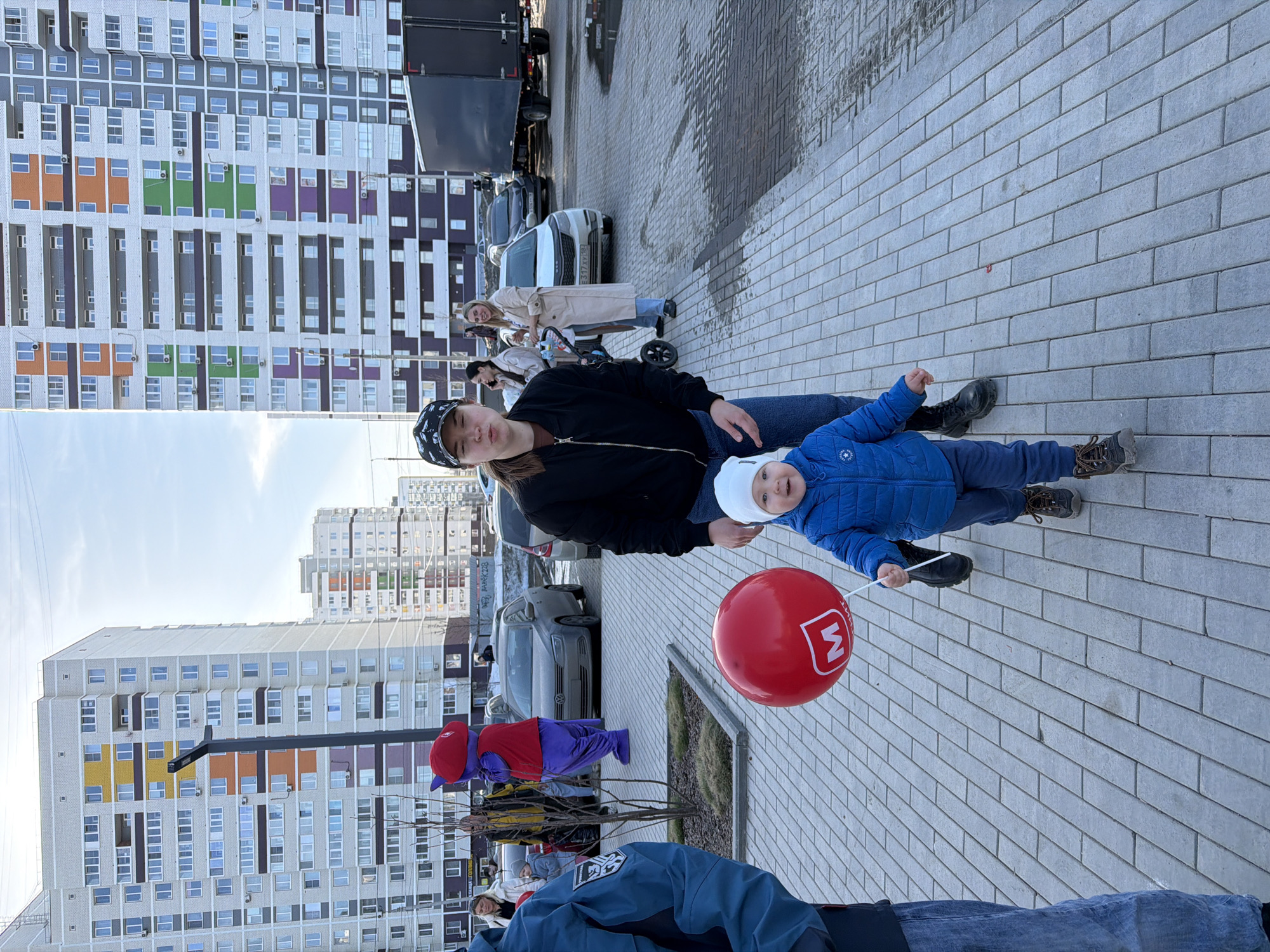 The image shows a street scene in Pervomaysky, Russia. A woman with a black jacket and patterned cap is seated on a paved area with a young child in a blue jacket and white hat on her lap. The child holds a red balloon with a white 'M' logo. In the background, a woman pushes a stroller. A person in a mascot costume stands among a small crowd. Multiple cars are parked nearby, and tall apartment buildings with colorful accents are visible. The sky is overcast.