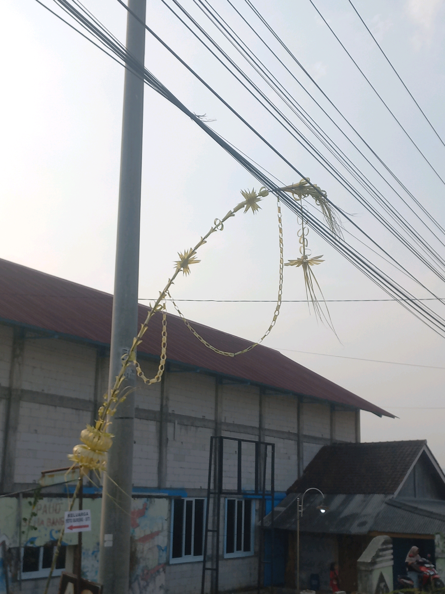 The image depicts a street scene in Baturetno, Indonesia, under a bright, hazy sky. A utility pole stands prominently in the foreground, adorned with yellow, palm-leaf-like decorations strung between its upper section and overhead wires. These decorations appear to be woven into intricate shapes resembling stars and chains. Below the utility pole, a large building with a dark red, sloped roof and white brick walls is visible. Some of its windows are covered with blue or black shutters. To the right of this building, a smaller, darker structure with a corrugated metal roof is partially visible, featuring a street lamp with a light on. In front of the larger building, there are some signs with text, though the text is not clearly legible. In the background, to the right of the smaller structure, two individuals are visible near a motorcycle. One person is seated on the motorcycle, and another is standing. The overall environment appears to be an urban or semi-urban setting with overhead power lines a significant feature of the landscape.