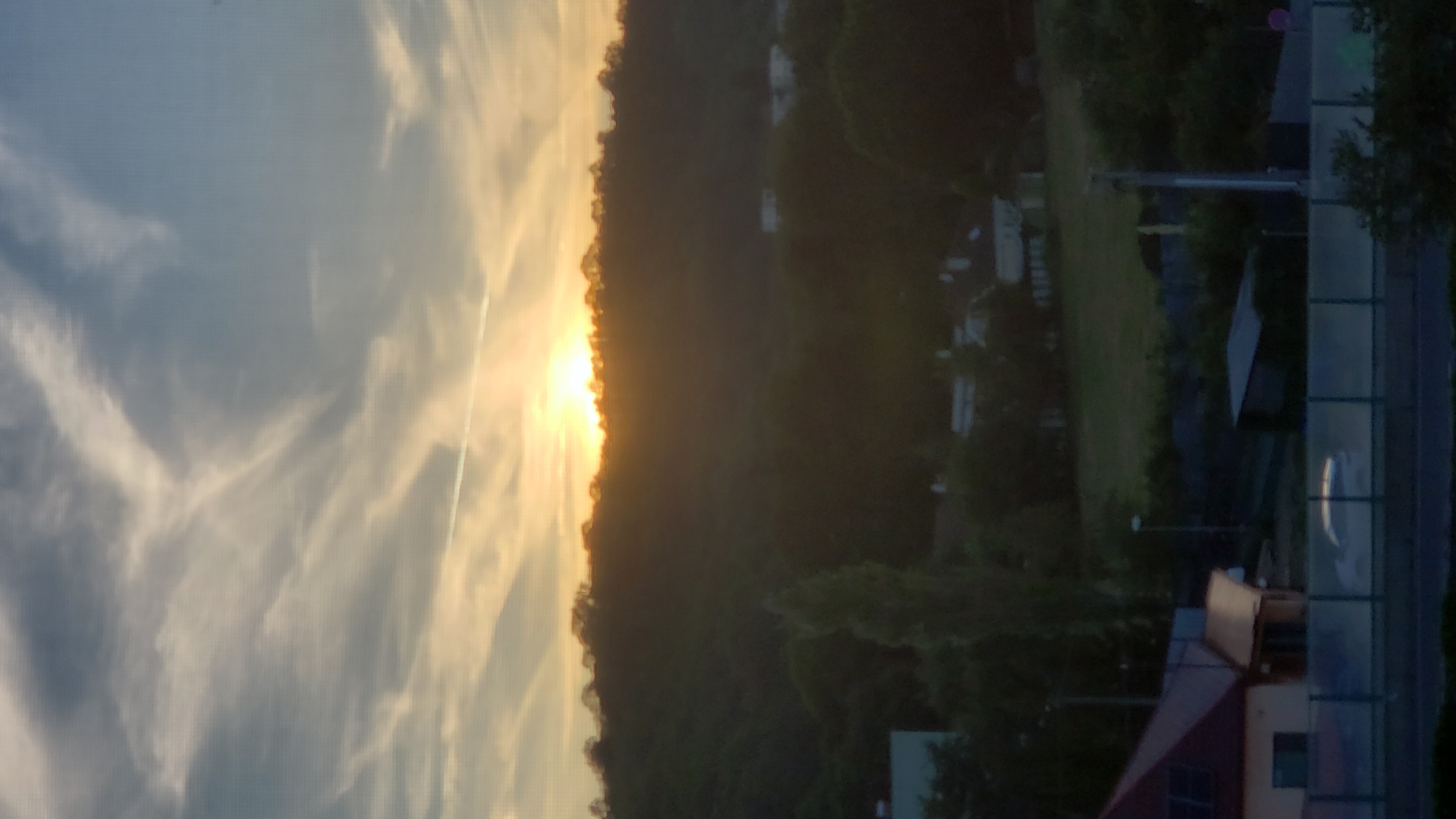 The image captures a twilight scene over a forested hill, with the setting sun casting a warm glow across the sky. Wispy clouds streak across the sky, some tinged with orange from the fading sunlight. A thin contrail from an aircraft is visible high in the sky. The foreground shows a residential area with houses, a road, and some trees. A white car is parked on the side of the road, its reflection visible on a transparent barrier. The overall ambiance is peaceful and serene.
This image does not provide enough information to determine the specific location within the city of Prešov, Slovakia.