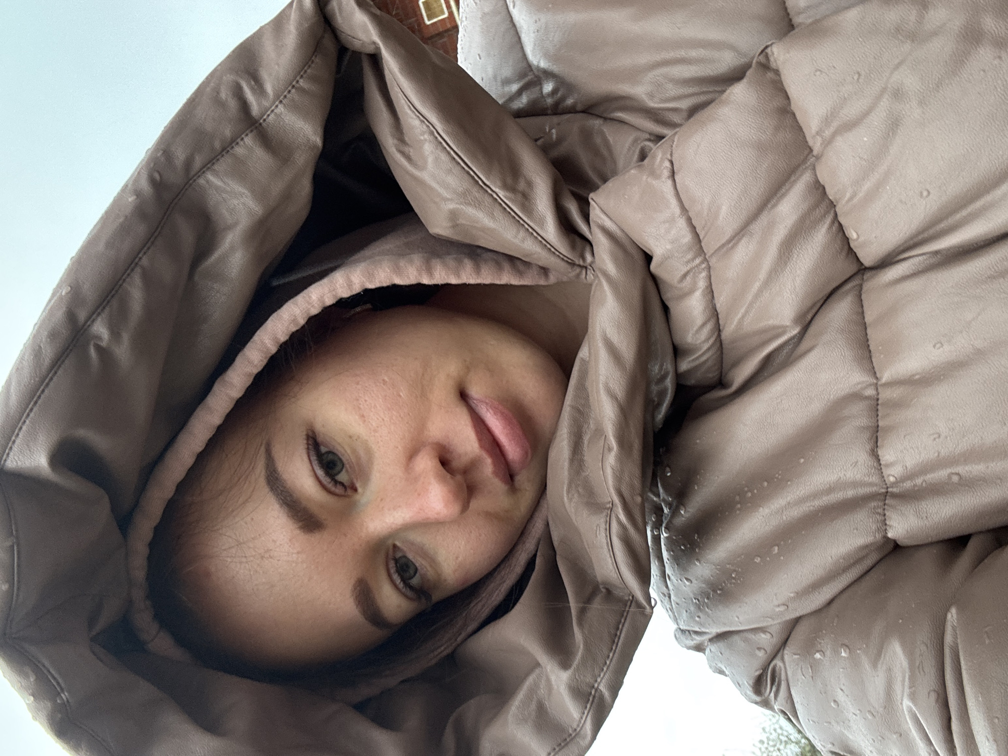 A woman in a brown hooded jacket poses for a selfie. The location appears to be outdoors in the city of Ферма, Russia. The jacket hood is up, partially obscuring her face. There are water droplets on the jacket, suggesting it may be raining or wet. The woman is looking directly at the camera. Her expression is neutral, with a slight smile and her tongue playfully sticking out.