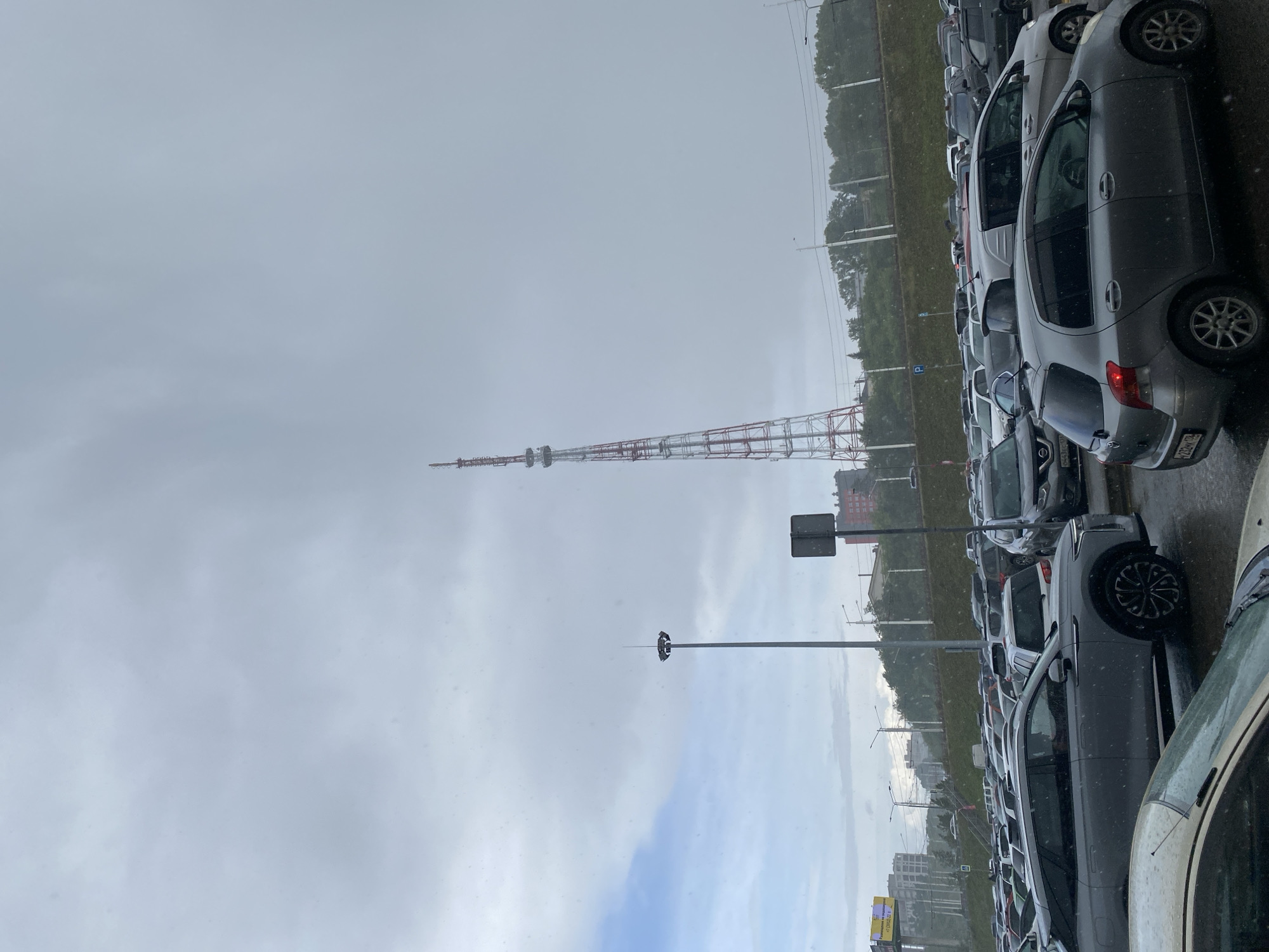 This image depicts a cloudy, overcast day in Irkutsk, Russia, with a large transmission tower visible in the distance. The foreground is dominated by a packed parking lot filled with numerous cars of various makes and models. The cars are parked in rows, indicating a public or commercial area, possibly a shopping center or transportation hub. The ground appears wet, suggesting recent or ongoing precipitation, which is also visible as white specks in the air, resembling snow or heavy rain. In the mid-ground, a tall, slender lamppost with multiple lights stands prominently, and a signpost is also visible. Beyond the parking lot, a grassy embankment lined with trees separates the immediate area from a city skyline featuring several buildings, including a taller, red-brick structure. The overall atmosphere is somber, consistent with the cloudy weather. There are no people clearly visible in the image.