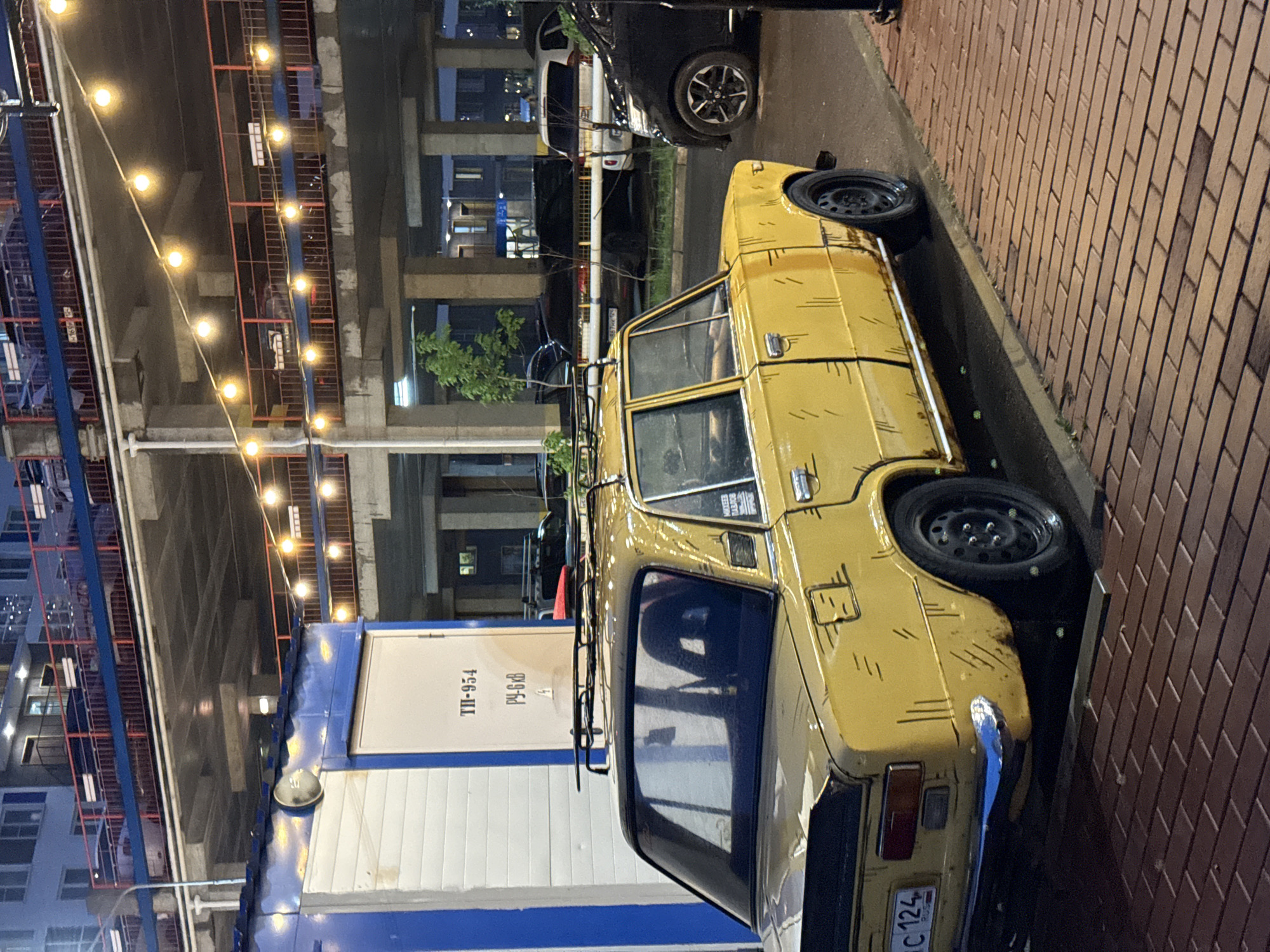 The photograph captures a yellow car parked on a paved surface in an urban environment. The vehicle is adjacent to a large blue and white container marked with the text "TI-954 Pч-6 ББ". Above the car, a bridge structure spans the scene, adorned with strings of illuminated lights. Behind the car, a building is visible, and other vehicles are parked nearby. The license plate of the yellow car reads "A C 124". Based on the text on the container, this location is in Krasnoyarsk, Russia. The overall scene is dimly lit, suggesting it may be night time.
