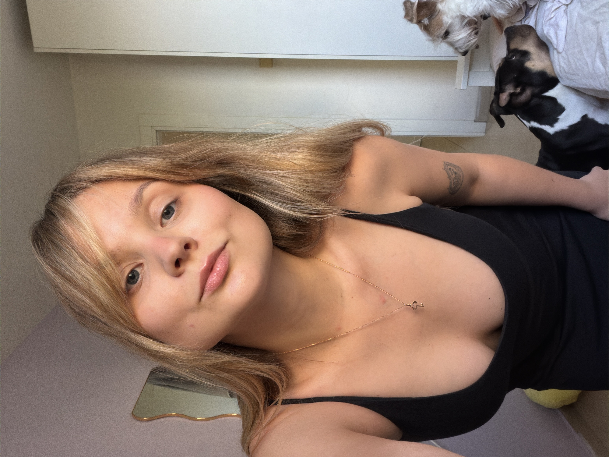 A selfie shows a young woman with long blonde hair and fair skin. She wears a black tank top and a delicate gold necklace with a small key pendant. Her lips are glossy, and her gaze is towards the camera. Behind her, there is a blurred background of white furniture and two dogs. One dog is white and furry, while the other is black and white. There is no information to identify a specific location in Tel-Aviv, Israel, or the occurrence of any action. There is an ornamental mirror visible as well.