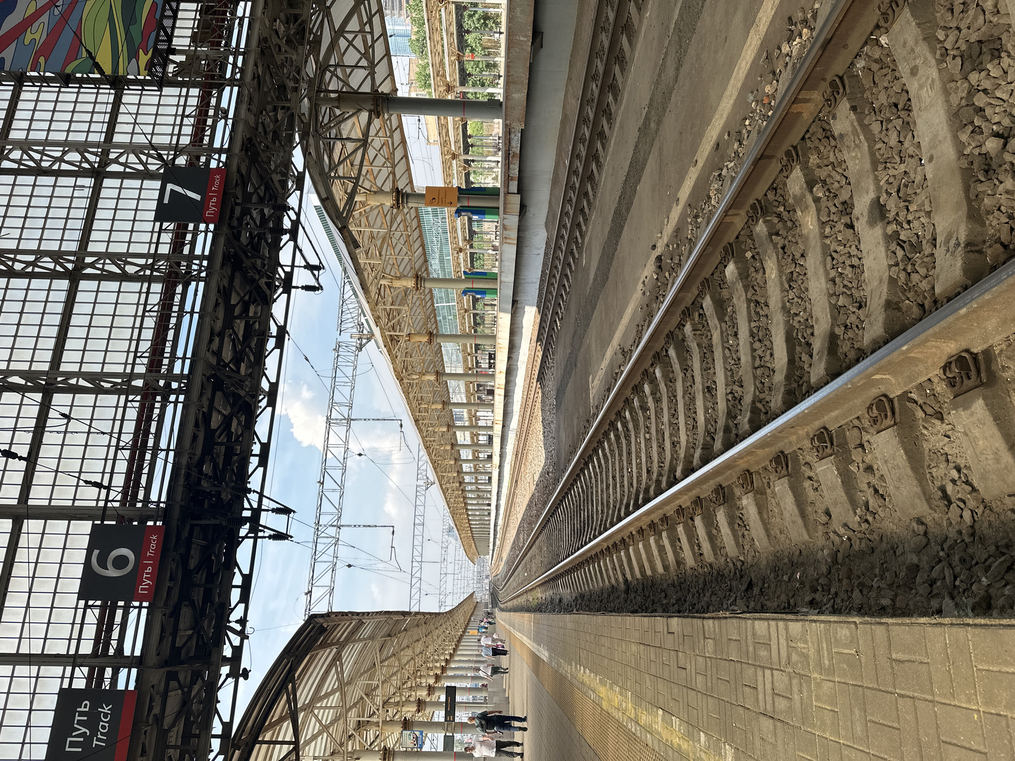 The image depicts a railway station platform in Moscow, Russia. The scene is framed by train tracks in the foreground, running towards a curved distance. Above the tracks, a covered platform with a transparent roof supported by numerous pillars extends into the background. Several people are visible walking on the platform to the left, dressed in casual attire. Signs indicating "Track 6" and "Track 7" are prominently displayed overhead, along with text in Russian and English that translates to "Path | Track." The environment suggests an active transportation hub with an emphasis on railway operations.