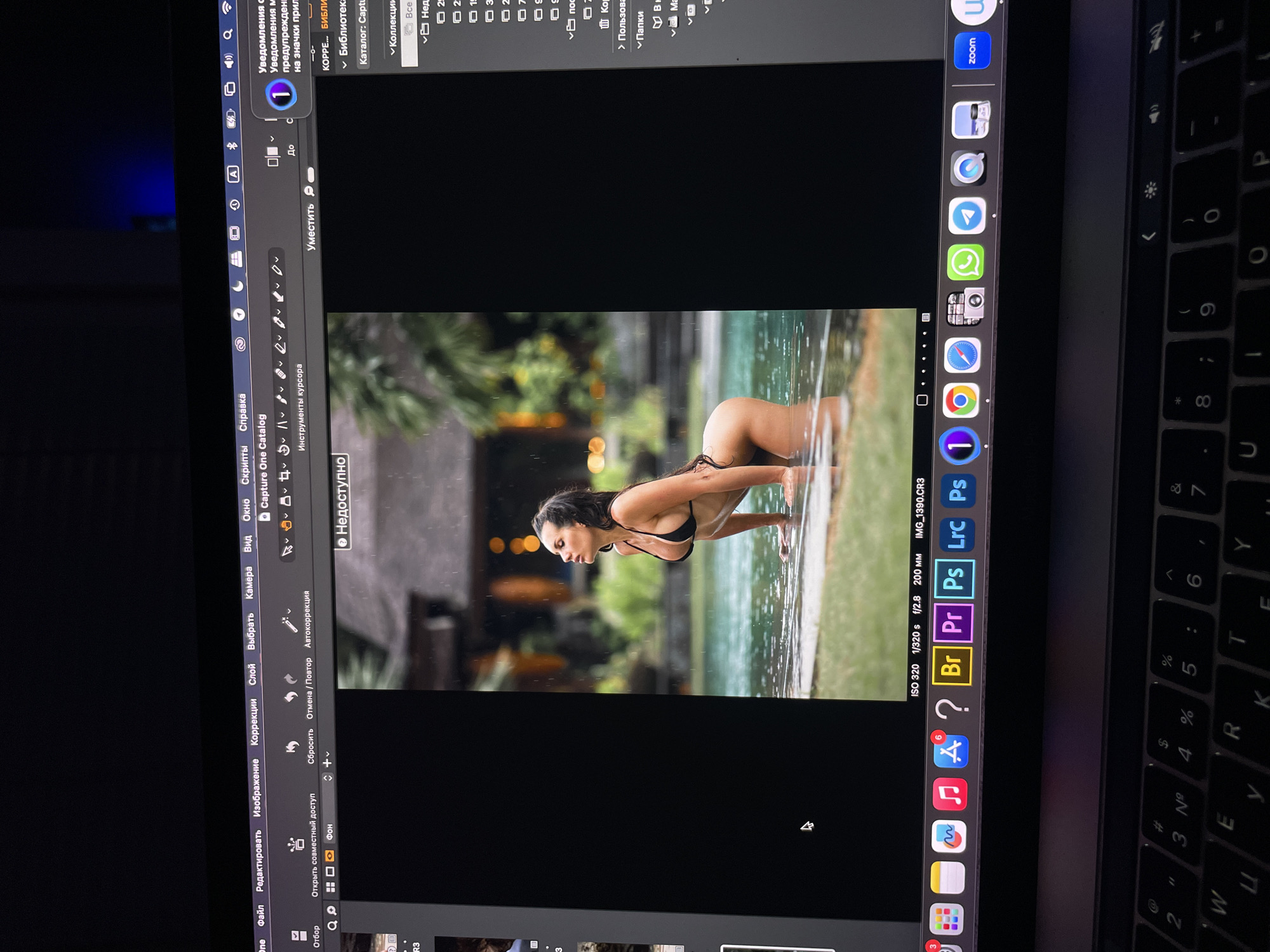 The image shows a laptop screen displaying a photo of a woman in a black bikini top leaning over a body of water. The woman has dark hair pulled back and is partially submerged in the water. Behind her, there are trees and blurred lights suggesting an outdoor setting, possibly a pool area or natural water source.
The laptop screen also displays the editing software Capture One with its various tools and menu options in Russian. Icons for other applications such as Photoshop, Lightroom, and a web browser are visible in the laptop's dock. The visible part of the keyboard shows some Cyrillic characters, likely belonging to the Russian keyboard layout.
There is no information about the location in the city of Kazan, Russia.