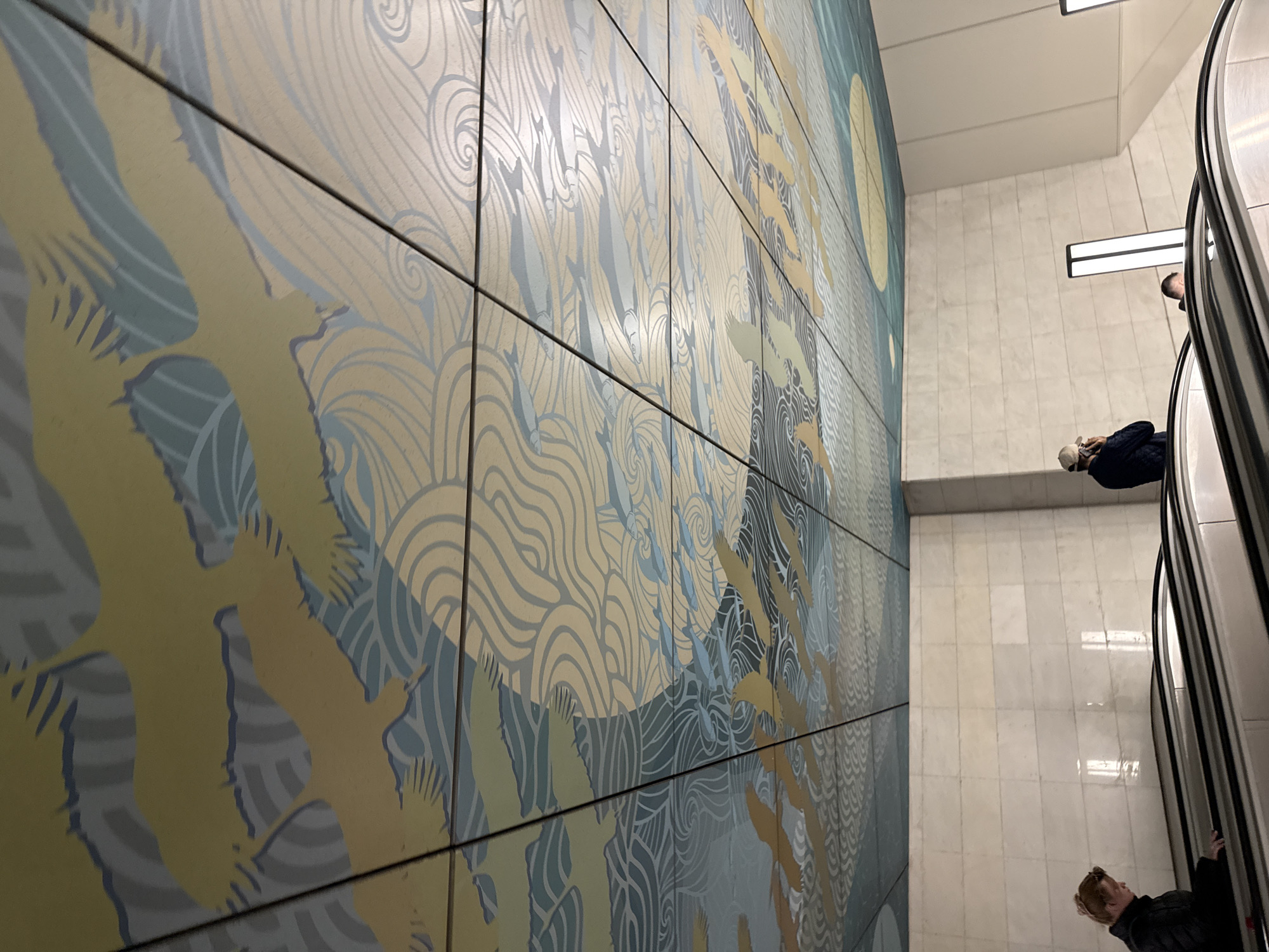A low-angle shot depicts a wall decorated with square tiles, featuring an image of flying birds and fish, adjacent to an escalator in the Moscow Metro, Russia. The wall mural is composed of a pattern with curving lines in shades of gold and blue-gray. Silhouetted figures can be seen riding the escalator. The environment includes white marble-like tiles on the walls and ceiling, illuminated by linear light fixtures.