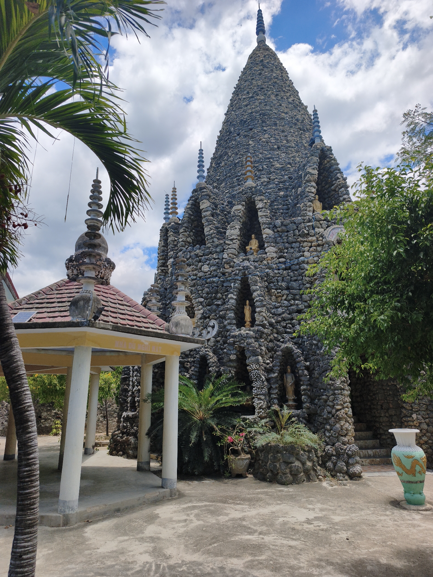 This image depicts the exterior of the Tu Van Pagoda in Cam Ranh, Vietnam. The pagoda is a unique architectural structure constructed primarily from a multitude of seashells and coral. Dominating the center is a large, conical tower adorned with numerous alcoves, many of which house small golden Buddha statues. Several smaller spires, also decorated with shells, punctuate the pagoda's design, resembling tiered stacks. To the left, a covered pavilion with a red-tiled roof and white pillars provides a contrasting, more conventional architectural element. Lush green foliage, including palm trees, surrounds the structures. A decorative vase with a dragon motif stands near the base of the main pagoda on the right. The ground is paved with concrete, and the sky above is partly cloudy.