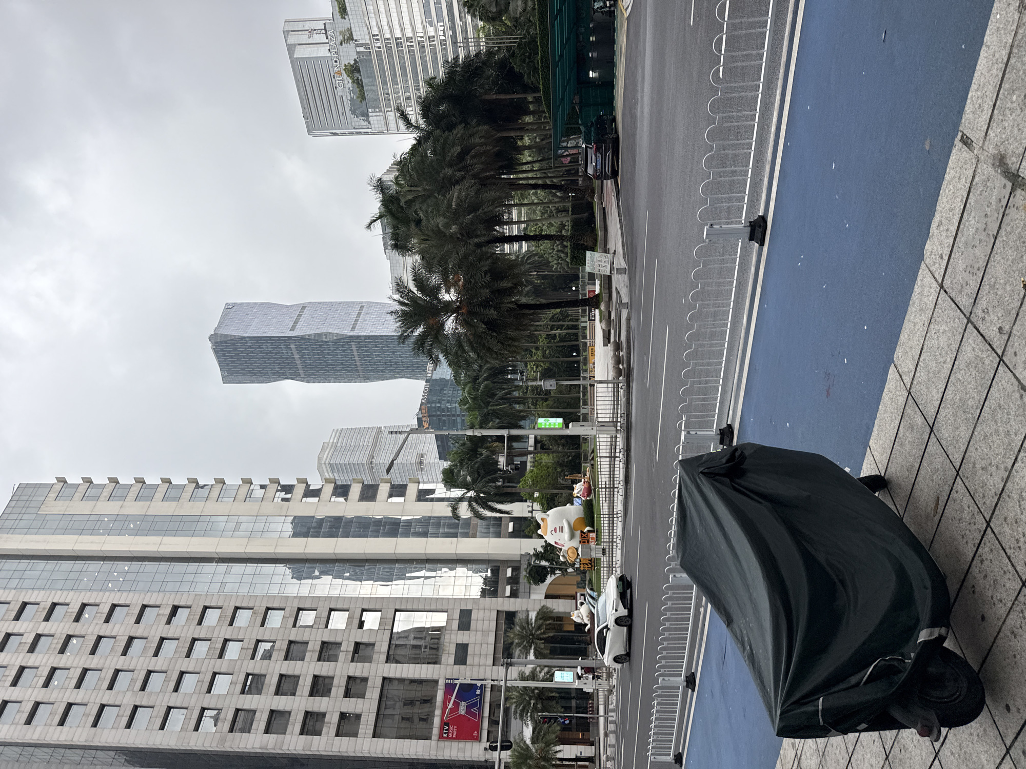 This image depicts a city street scene in Tianhe District, China, on an overcast day. The foreground features a motorcycle covered with a dark fabric tarp, resting on a sidewalk with a patterned stone surface. Adjacent to the sidewalk is a bright blue lane, possibly for bicycles or pedestrians, separated from the road by a white metal fence.
The street is paved and appears to be a multi-lane road with white lane markings. Several cars are visible on the road, including a white car in the middle ground. The scene is lined with palm trees, creating a tropical or subtropical ambiance.
In the background, towering modern skyscrapers dominate the skyline. One prominent building has a distinctive angular design. Other buildings are characterized by glass facades, reflecting the cloudy sky. Some text is visible on a billboard on a building in the lower left, which appears to advertise "KTV MUSIC". There are also some decorative animal statues on a grassy area near the buildings, one of which resembles a large white cat. The overall mood is subdued, likely due to the overcast weather.
