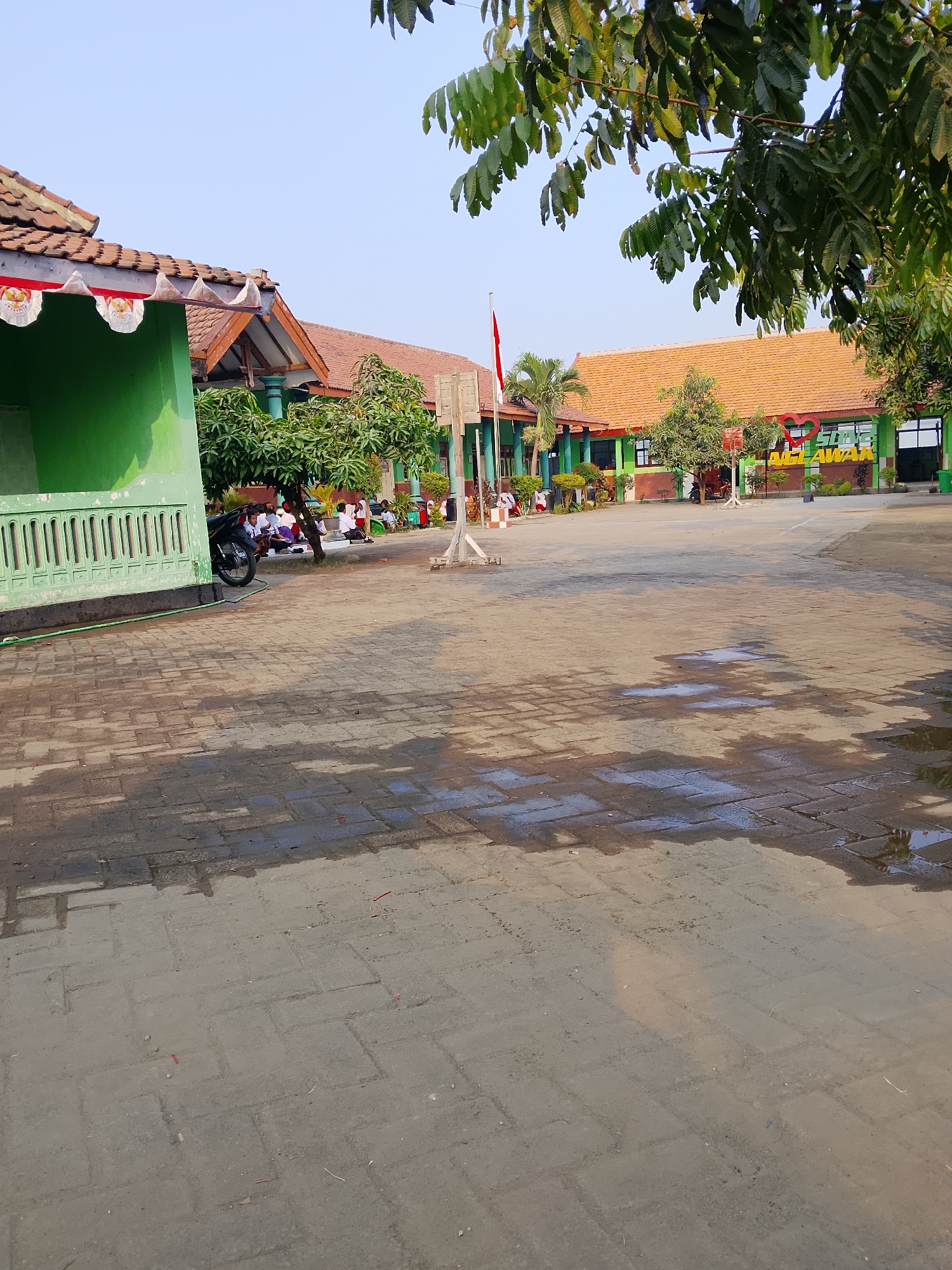 The image depicts an outdoor scene, likely a schoolyard, in the city of Kepuh, Indonesia. The ground is paved with interlocking bricks, with several puddles reflecting the sky. In the foreground, the paved area extends towards the background, showing some debris scattered on the ground.
To the left, a green building with white decorative railings is visible. A parked motorcycle is positioned next to this building. Underneath a tree, a group of children in white uniforms are sitting on the ground.
In the background, several school buildings with terracotta tile roofs are arranged around a central courtyard. A flagpole with an Indonesian flag is prominent in the courtyard. More trees and students can be seen throughout the courtyard. A sign with the words "SD N 1 AGELAWAR" and a heart symbol is mounted on a wall. The overall environment appears to be a sunny day with a clear blue sky.