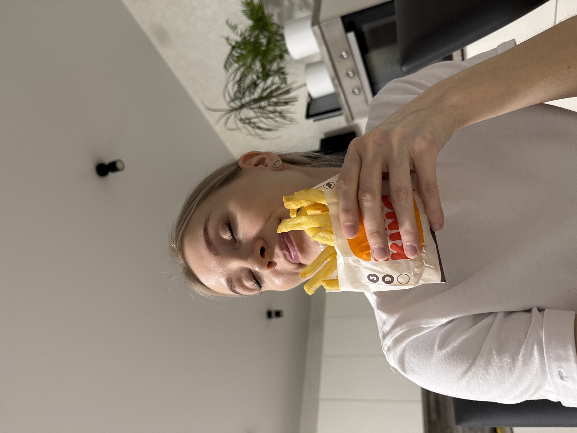 A woman is holding a bag of fries. The bag appears to be from Burger King. The woman is fair-skinned with blonde hair. She is wearing a white shirt. In the background, there is a plant and what appears to be a microwave or oven. The location is implied to be Izhevsk, Russia.