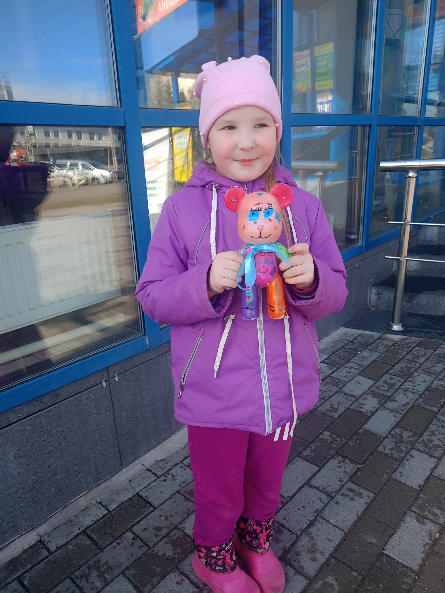 A young girl stands outside a building in an urban setting. She is wearing a pink hat with small white beads, a purple winter coat, and matching purple pants. Her pink boots have a pattern of hearts and flowers. She is holding a handmade doll with a pink bear head adorned with painted facial features and a blue ribbon tied around its neck. The doll's body consists of colorful tubes. The building behind her has a blue window facade reflecting the street scene, including cars and other buildings. The ground is paved with gray and black bricks, some of which appear wet. A silver handrail is visible to the right of the girl.
