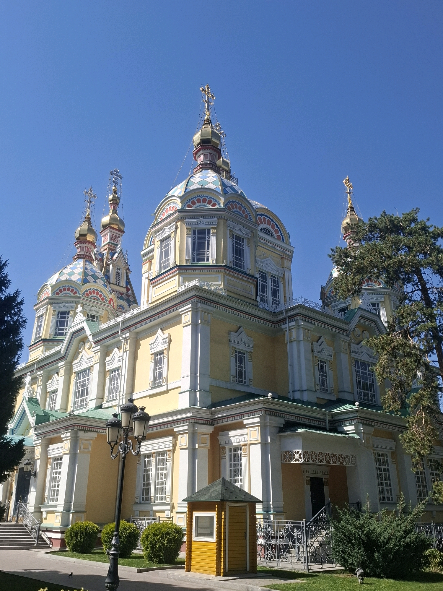 The image depicts the Ascension Cathedral (Zenkov Cathedral) in Panfilov Park, Almaty, Kazakhstan, under a clear blue sky. The cathedral, a prominent landmark, is a vibrantly colored, multi-domed Orthodox church. Its exterior features a pale yellow facade with white decorative elements, and several gold-topped domes. Some domes are adorned with colorful geometric patterns in red, blue, and white. The architecture is intricate, with arched windows and ornate detailing visible on the facade.
In the foreground, a tall black lamppost with three lights stands on the right side of a paved pathway. To the left of the lamppost are neatly trimmed green bushes. Adjacent to the bushes and further right, a small, bright yellow wooden structure with a window is situated. Beyond this structure, a black metal fence with decorative scrollwork encloses a grassy area. A large evergreen tree with dark green foliage is visible on the right side of the cathedral, partially obscuring one of its domes. Steps lead up to an entrance of the cathedral, and a black metal railing lines the steps. A black bird is visible on the grass in the lower left of the frame. The overall scene is one of a well-maintained park surrounding a significant religious building.
